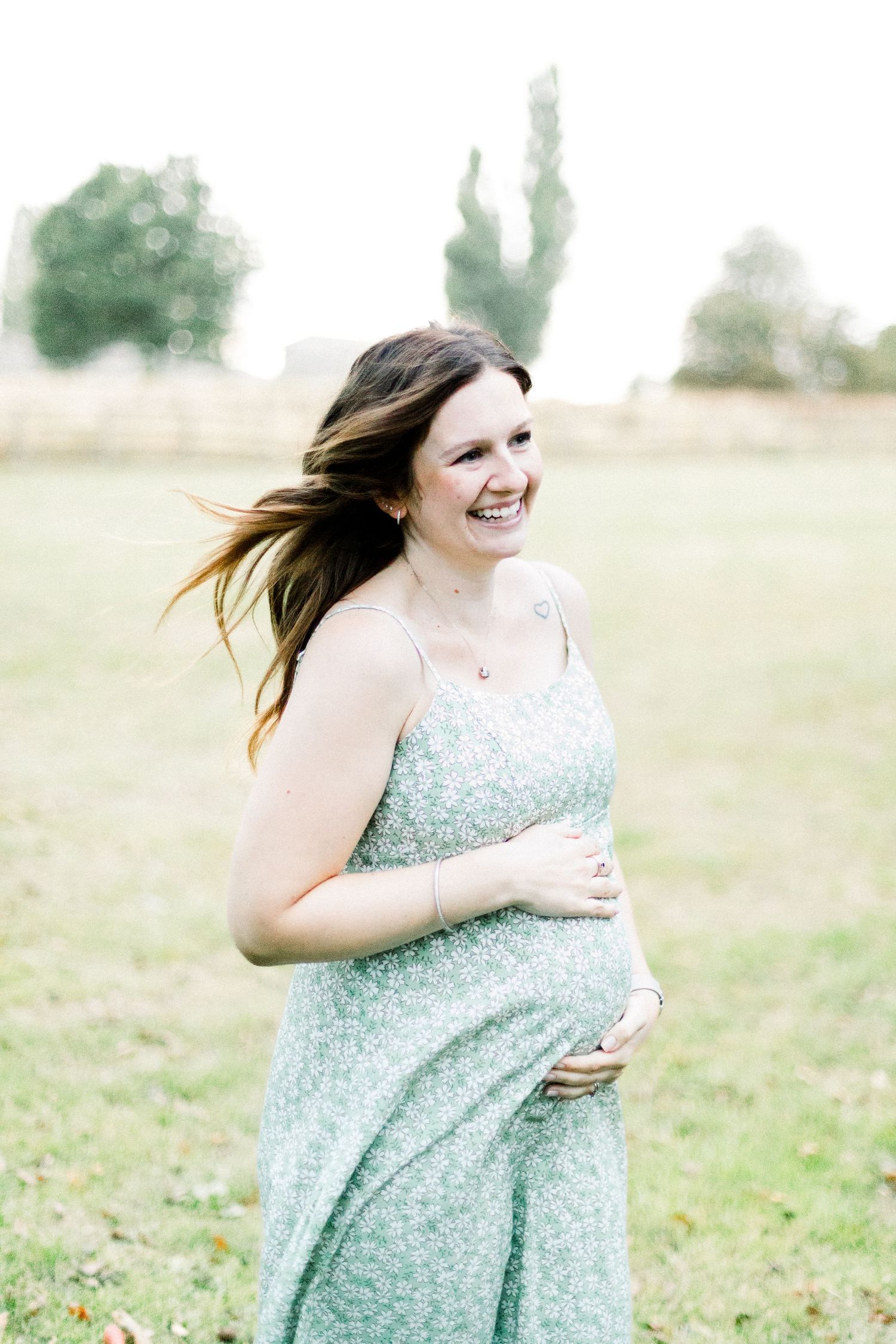 Smiling pregnant woman in floral mint dress cradling baby bump outdoors in green field with trees.