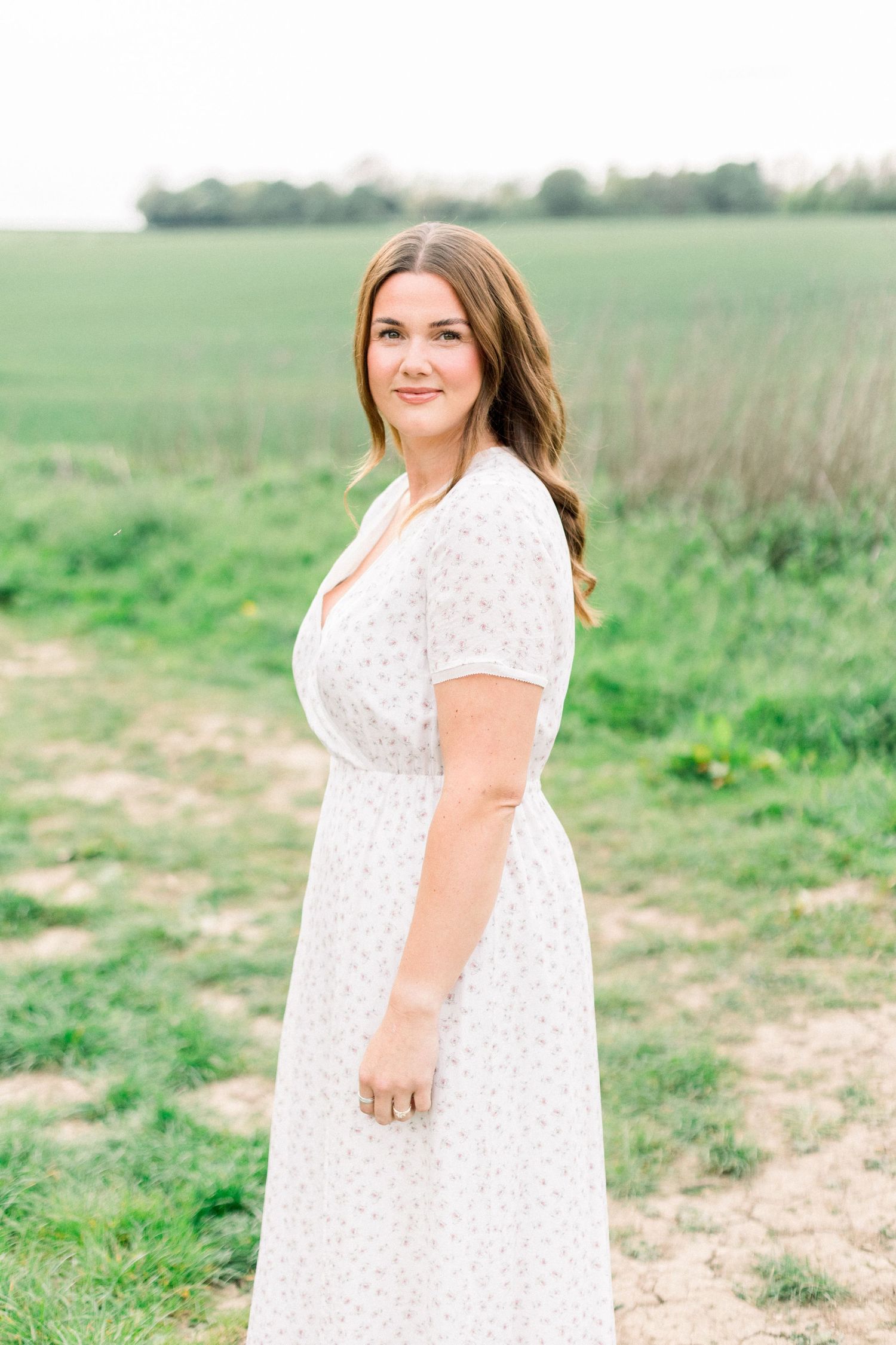 Young woman in white floral dress poses on a rural dirt path beside a lush green field.