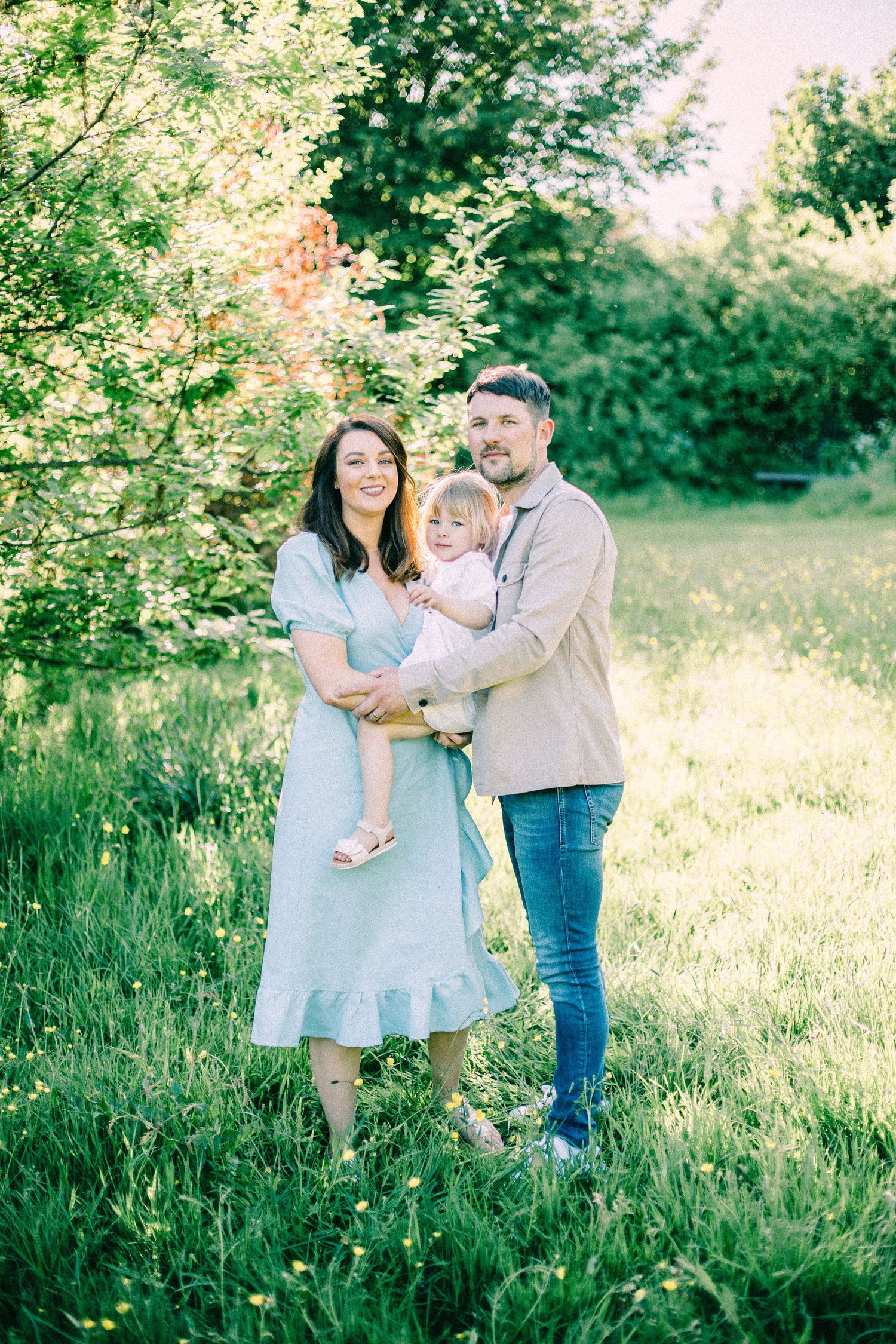 A smiling woman in a light blue dress and a man in a beige shirt hold a toddler in a sunny green meadow.