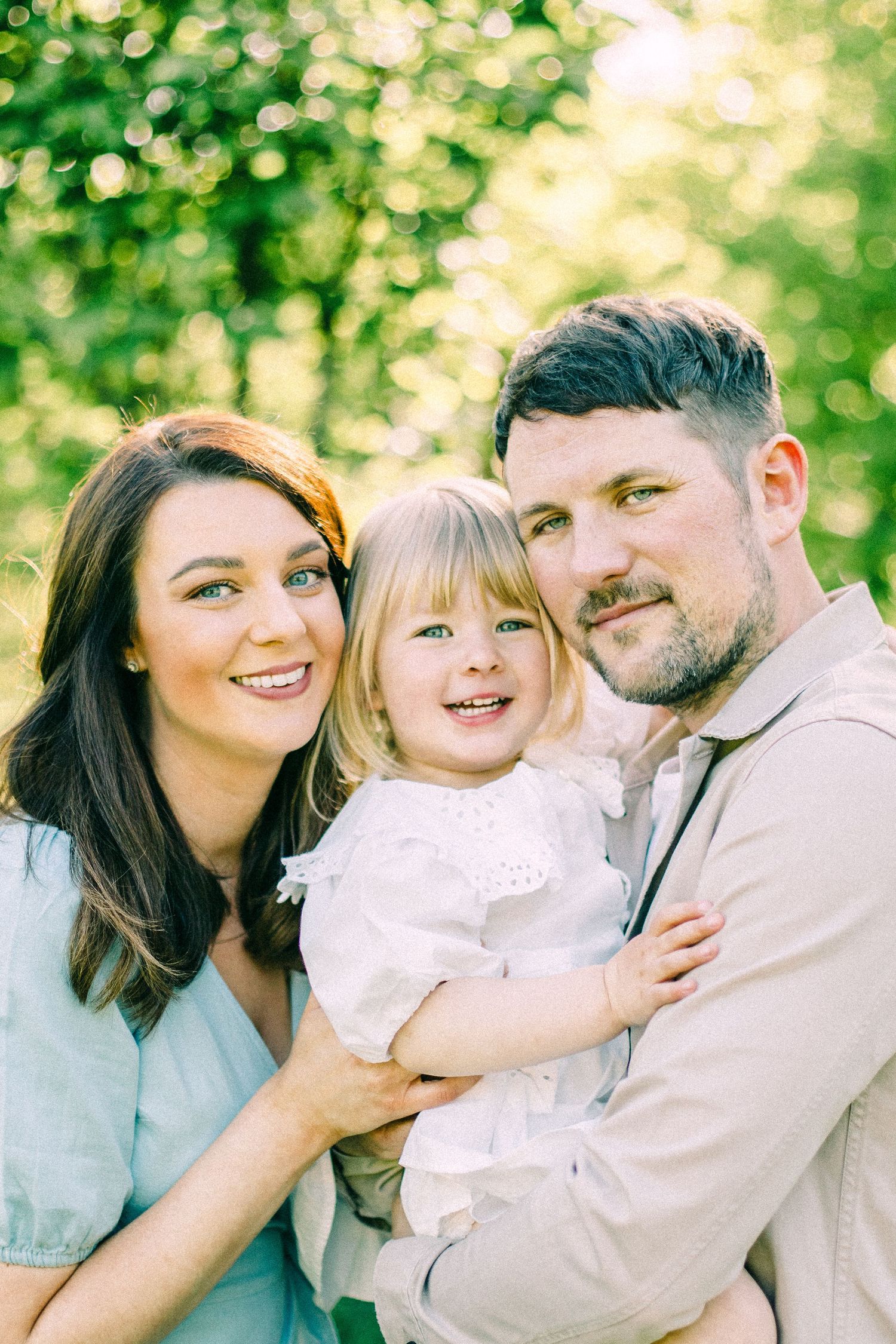 Smiling woman and bearded man hold a happy blonde toddler in a white dress outdoors, with sunlit green trees softly blurred behind them.