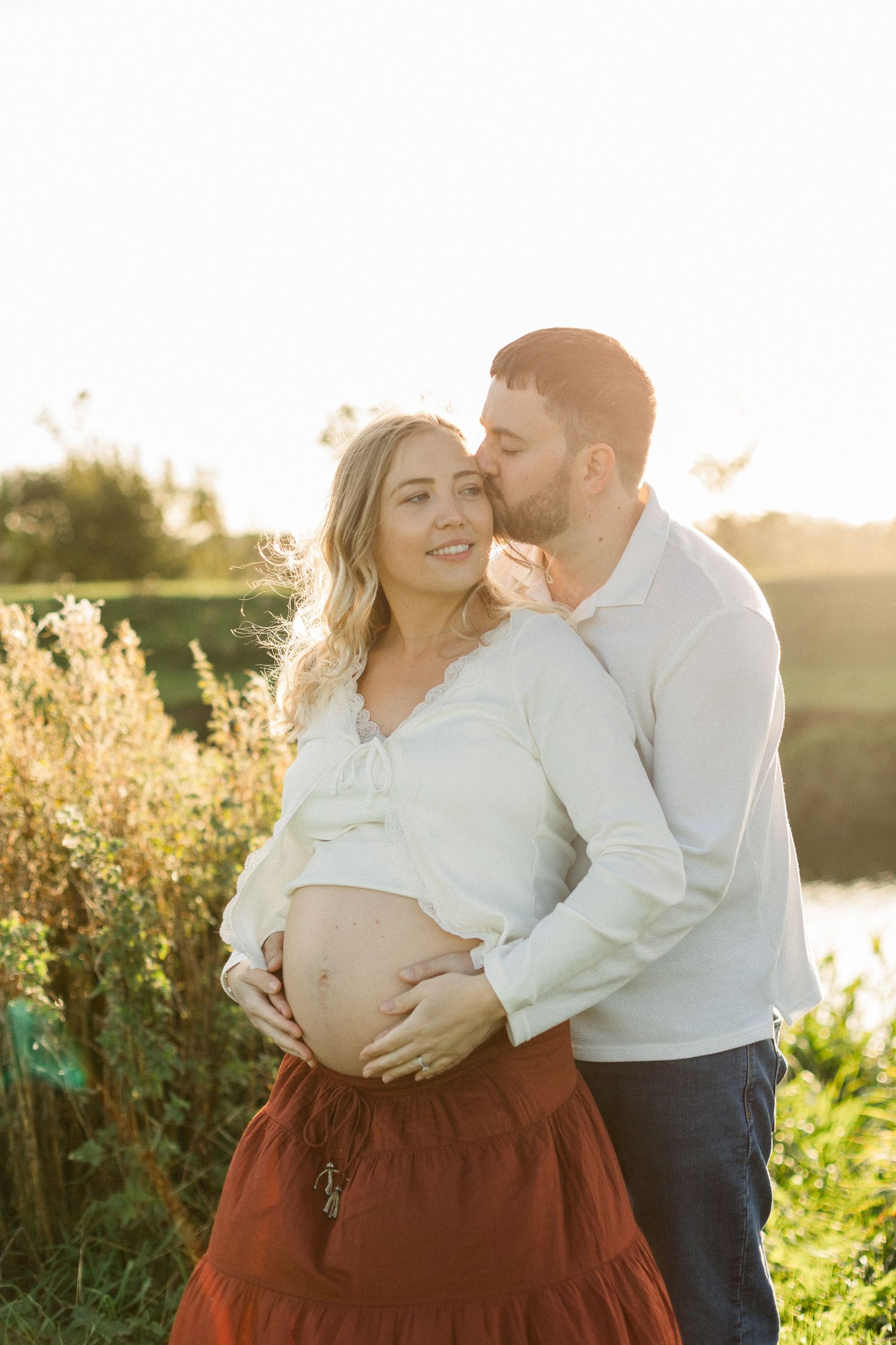 A man kisses a smiling pregnant woman outdoors at golden hour, both cradling her baby bump in a sunlit field.