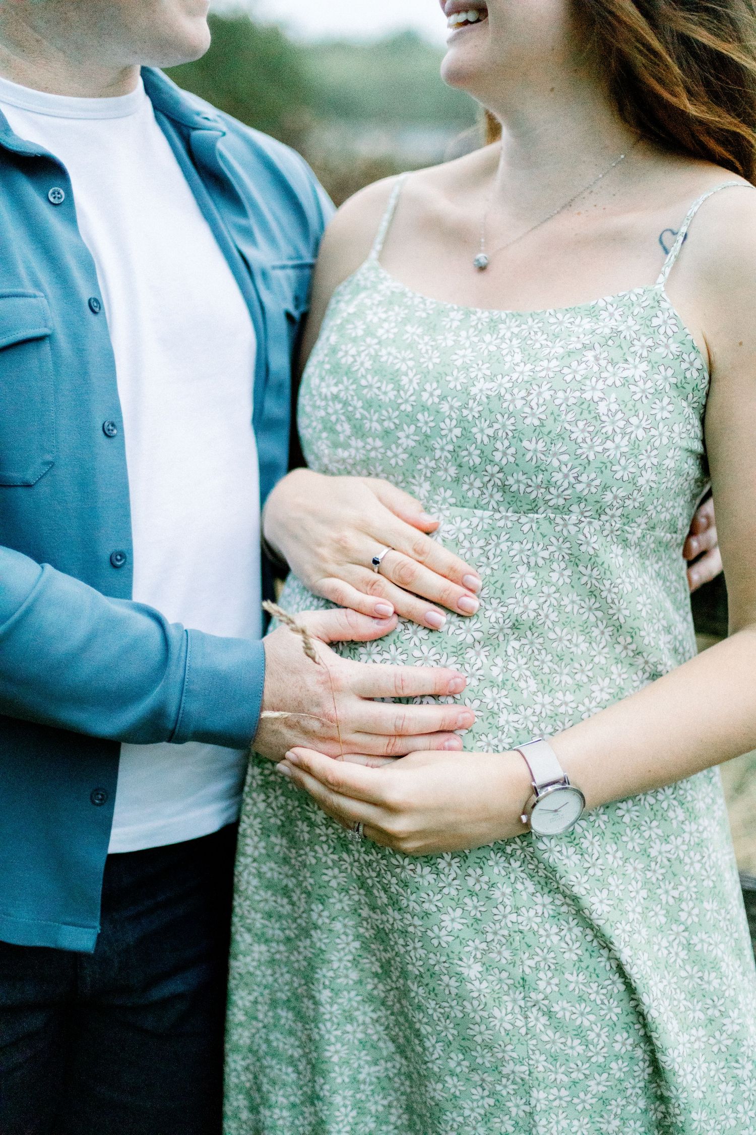 Couple cradling pregnant belly together, woman in floral green dress and man in blue shirt, maternity photo outdoors.
