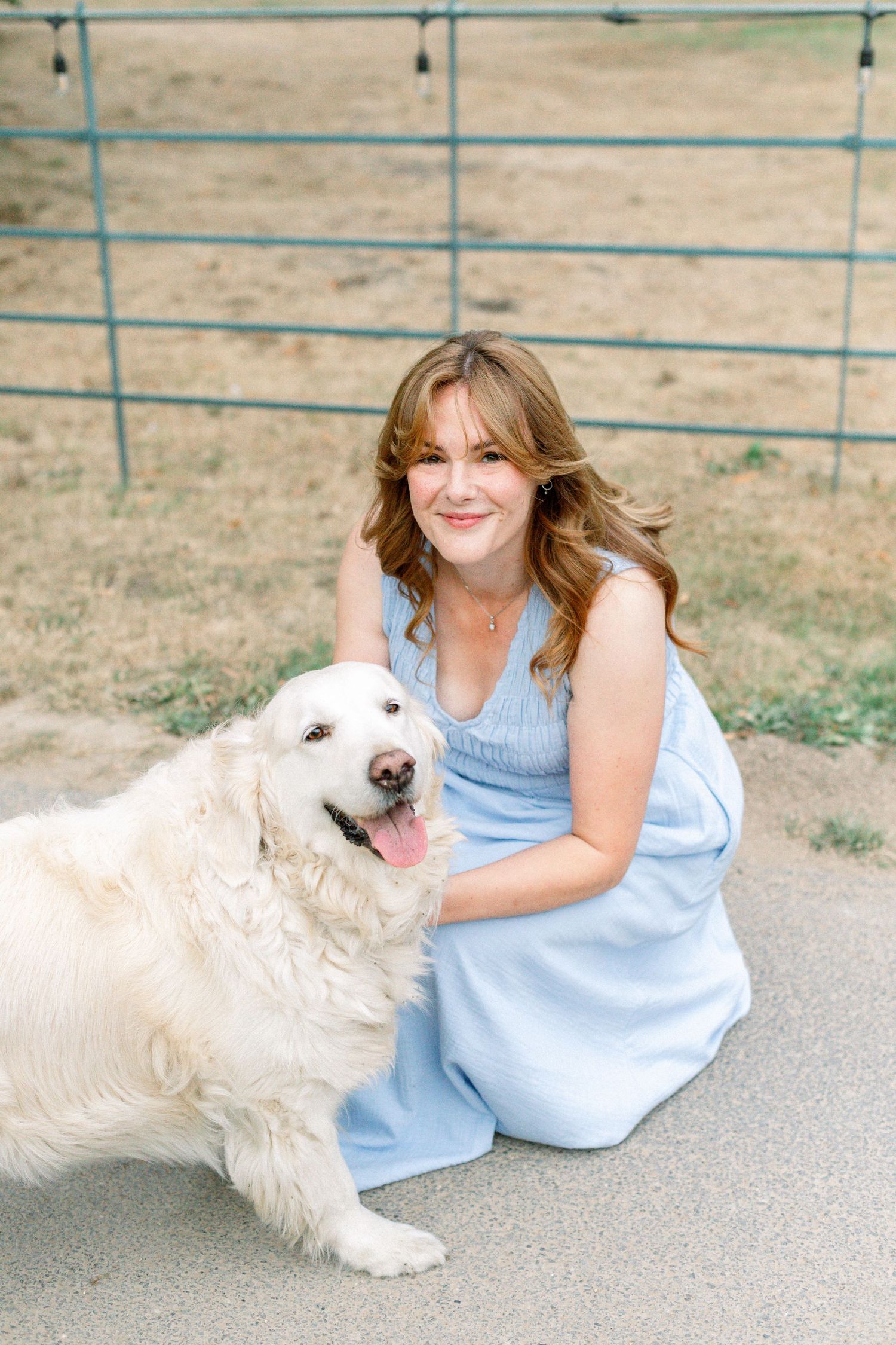Woman in blue dress smiling and posing with a fluffy white dog outdoors near a fence.