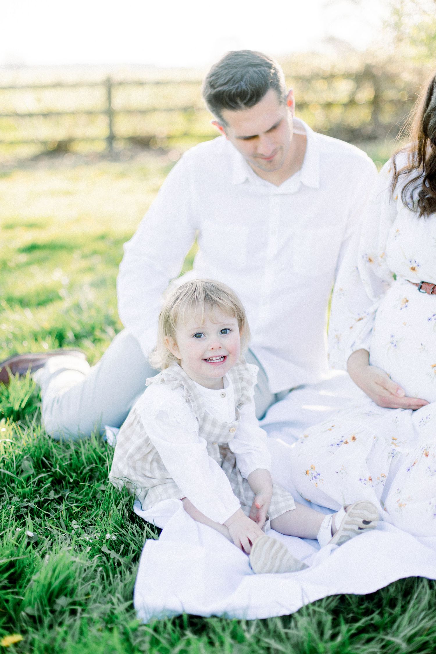 Family wearing white clothing sits together on grass during golden hour outdoor photoshoot.
