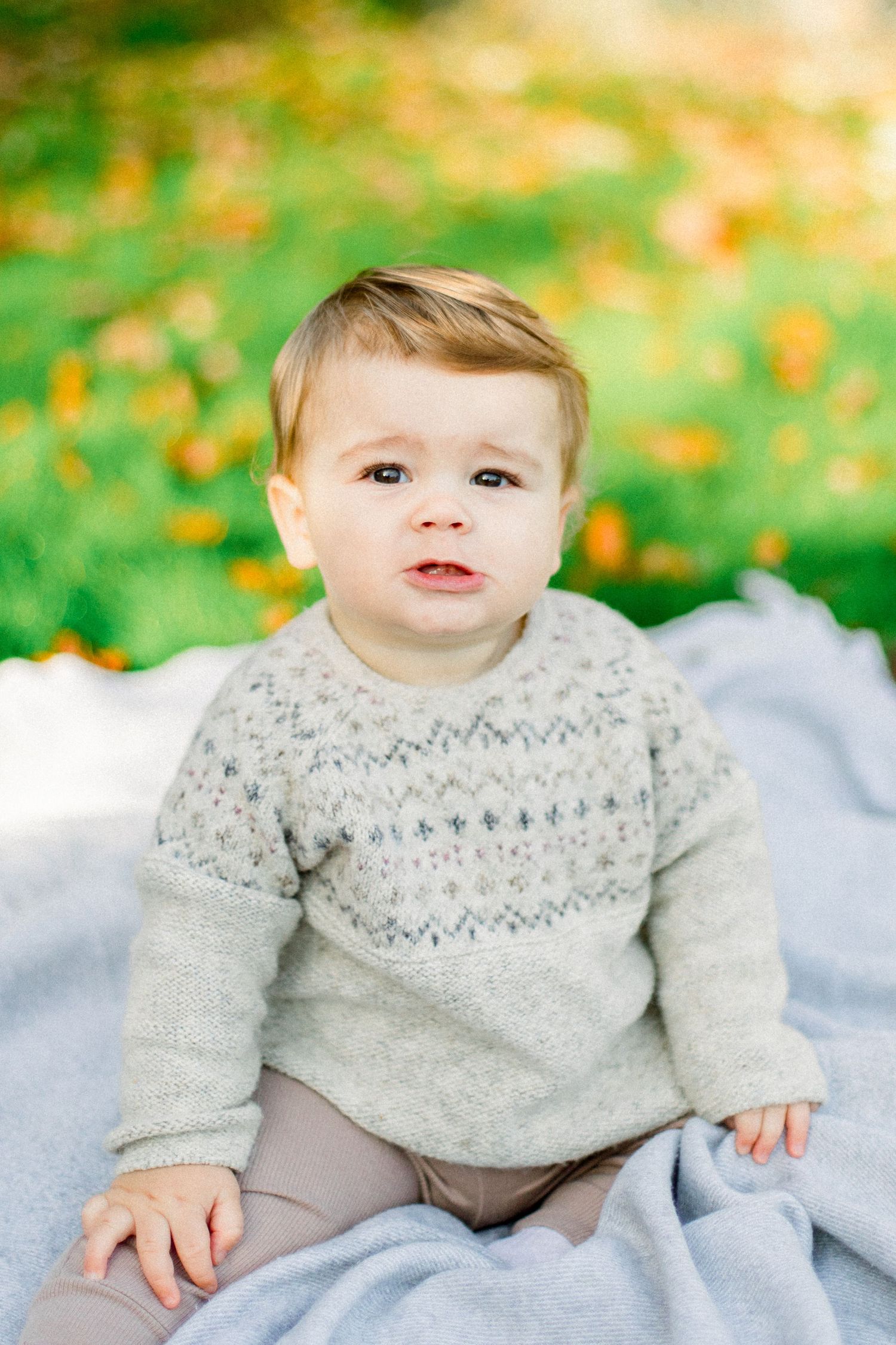 A small child wearing a grey patterned sweater sits on a light blue blanket outdoors with autumn leaves in background.