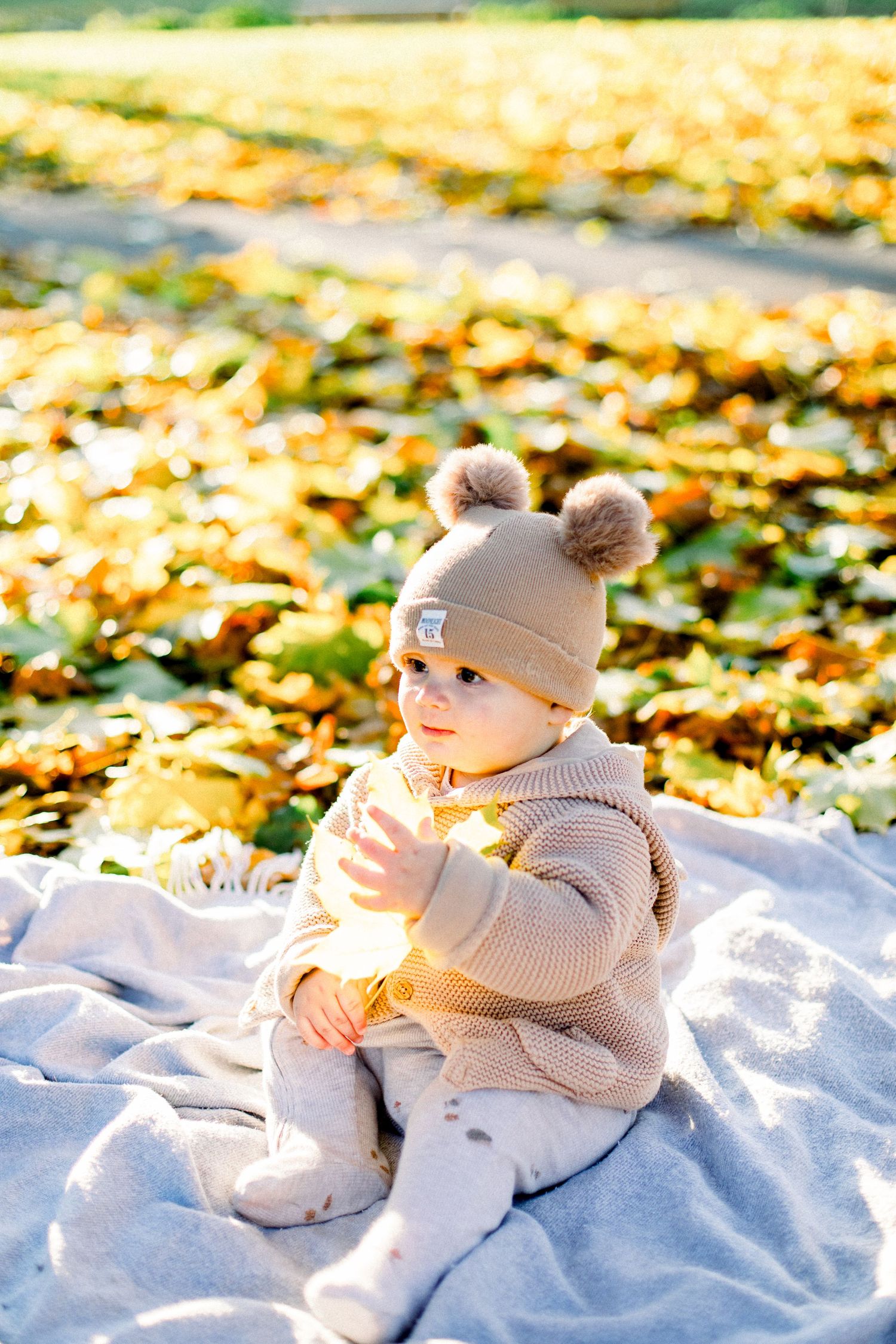 Adorable baby in beige knit outfit and pom-pom hat sitting on blanket surrounded by golden autumn leaves.