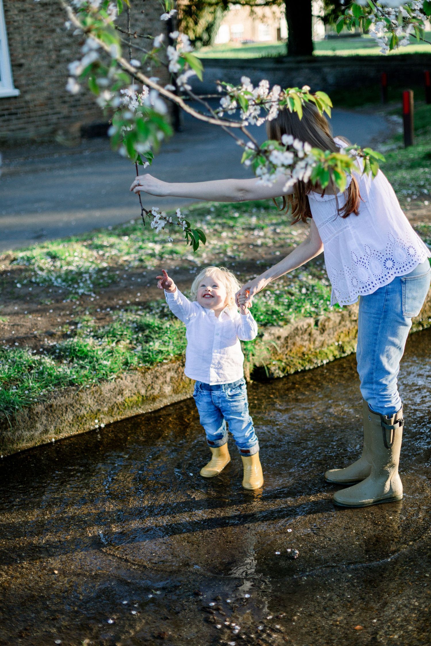 Mother and toddler playing under blooming cherry tree in spring, both wearing jeans and rubber boots near a puddle.