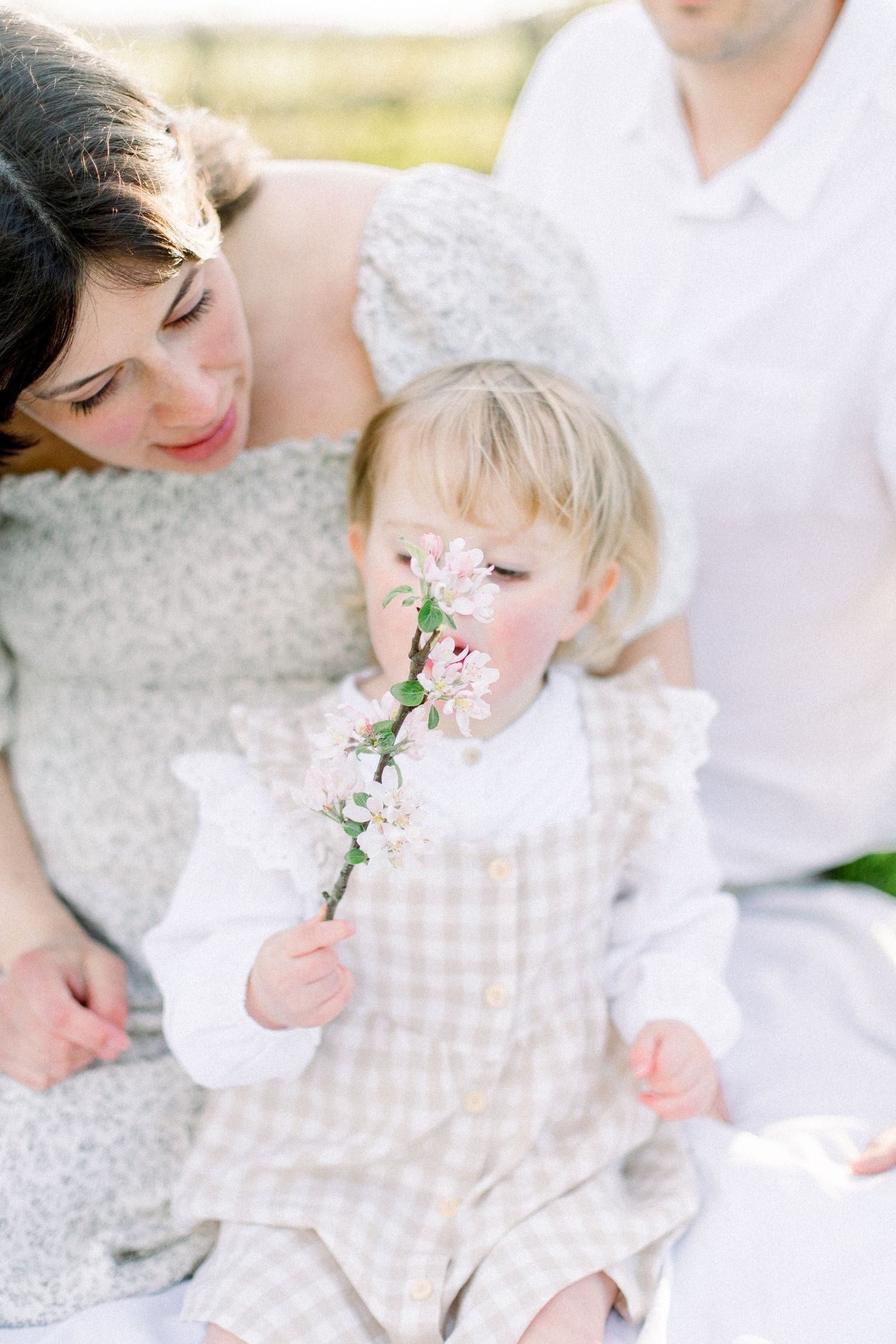 A loving mother watches her blonde toddler daughter smell a delicate branch of pink spring blossoms outside.