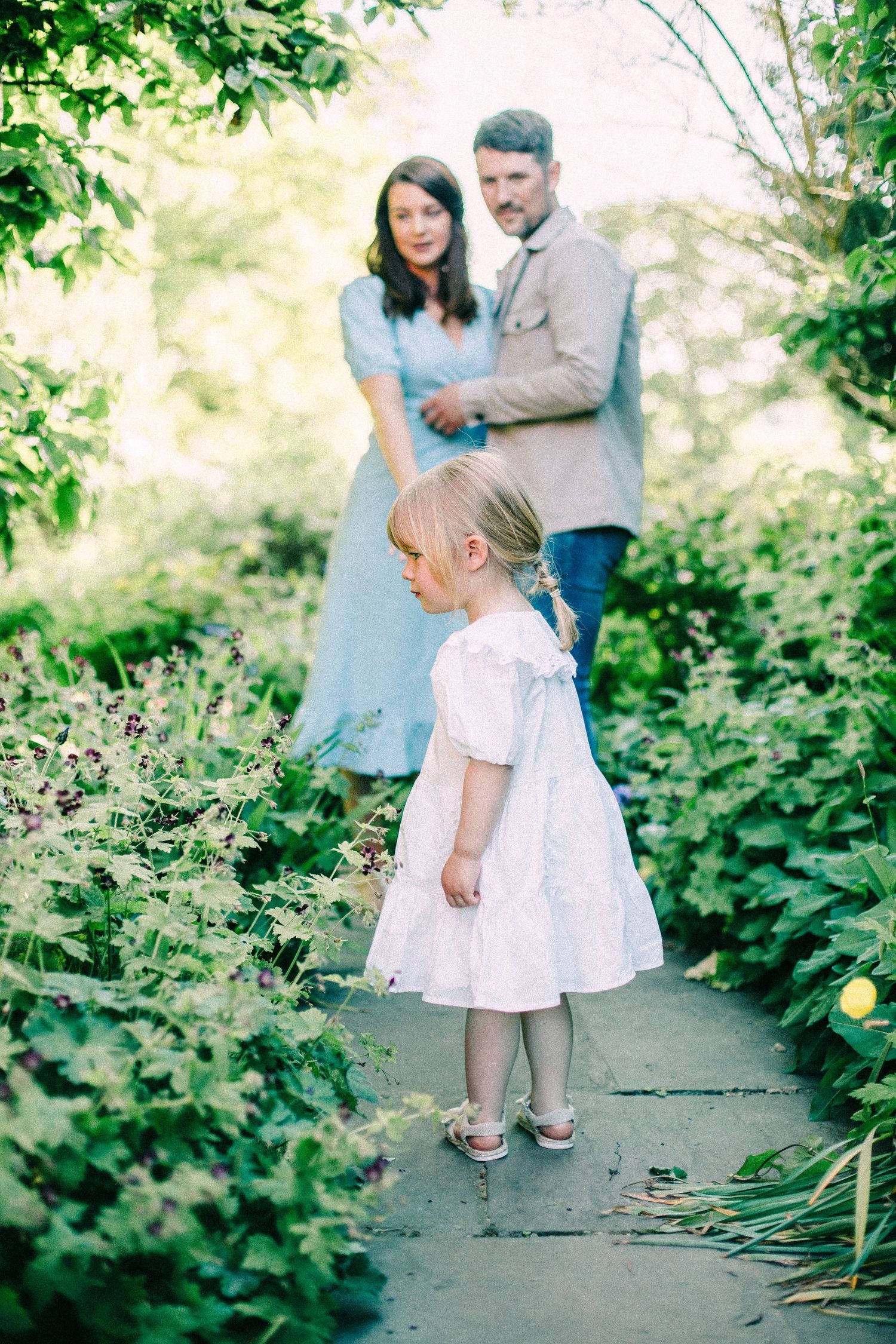Young girl in a white dress walks along a stone garden path while a man and woman stand together among lush green plants behind her.