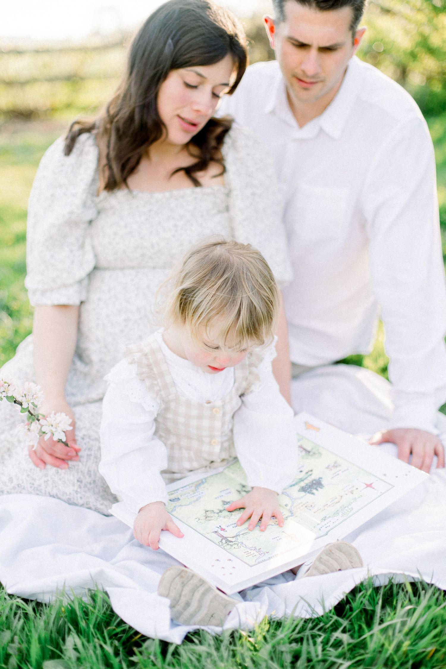 A young family sits together on a white blanket outdoors as their toddler daughter explores a picture book.