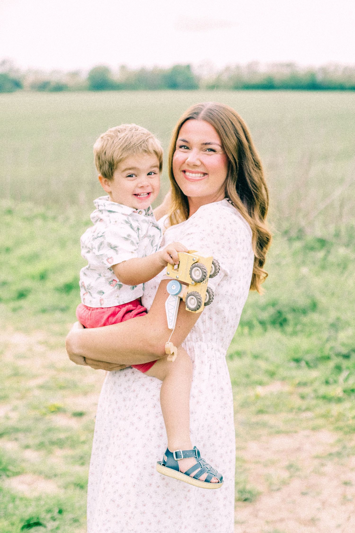 Smiling woman in a floral dress holds a happy toddler holding a toy truck while standing in a green grassy field.