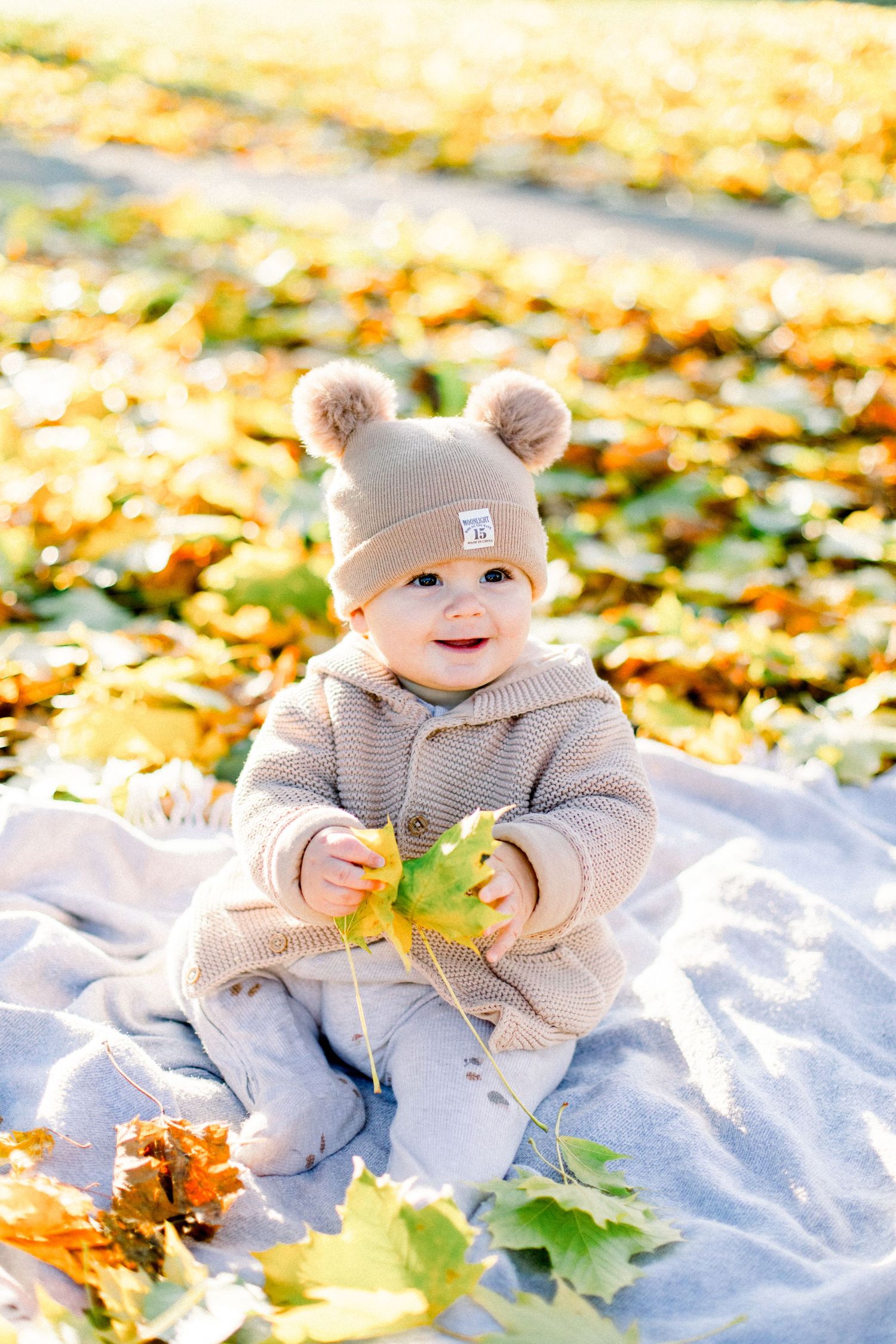 Baby in winter wear playing with fallen leaves on a white blanket during autumn.