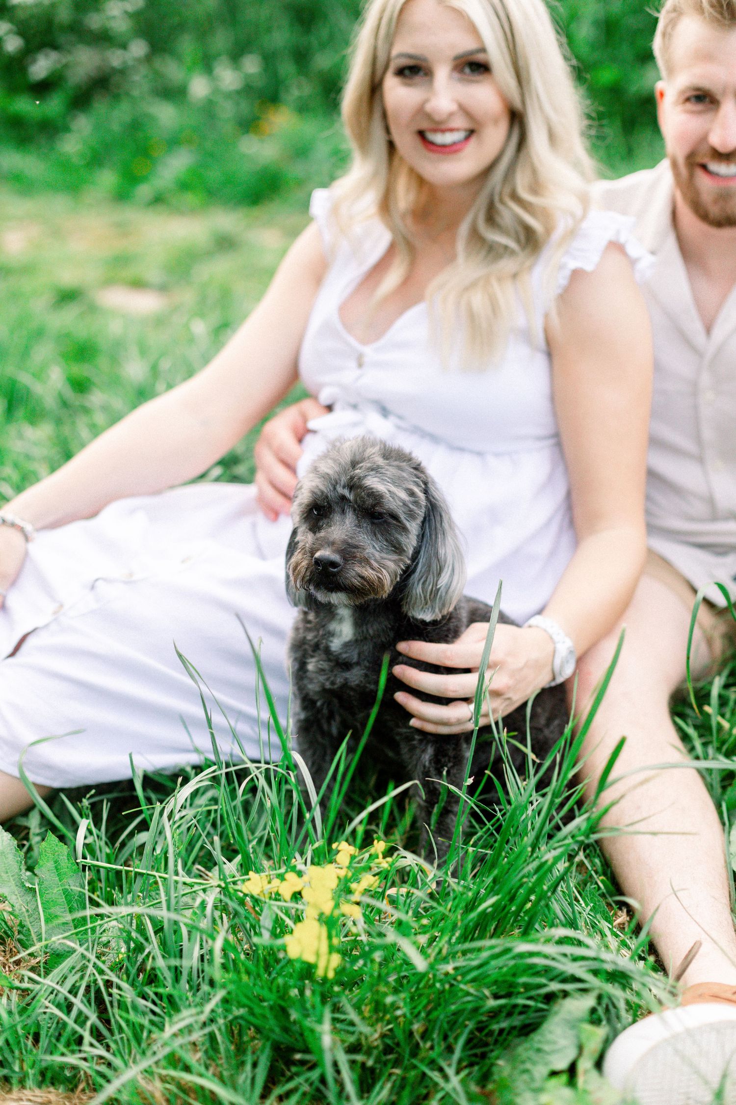 A smiling couple in white outfits sit in green grass outdoors, holding a small black fluffy dog near yellow wildflowers.