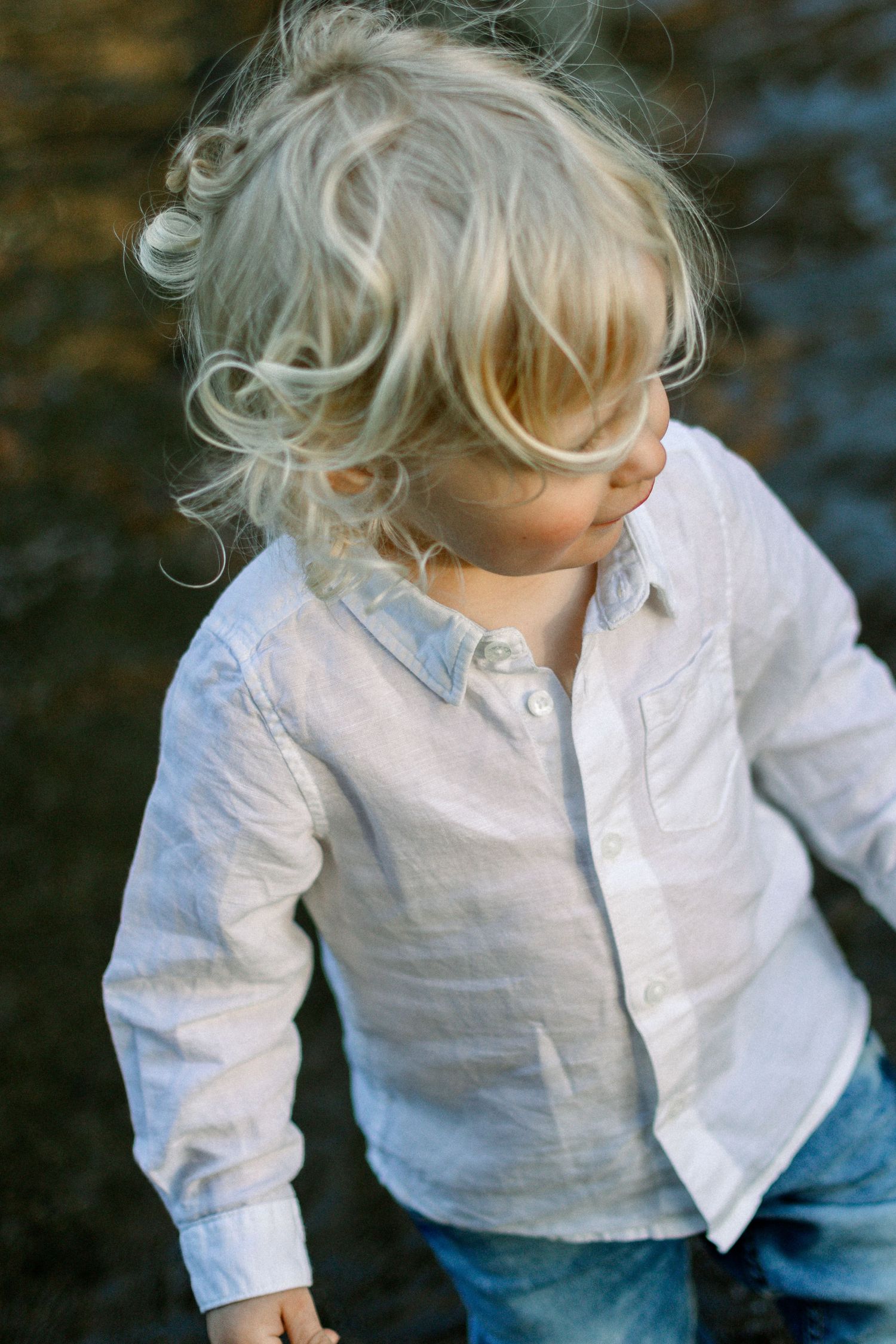 Blonde toddler in white linen shirt and jeans looking down outdoors in natural light.