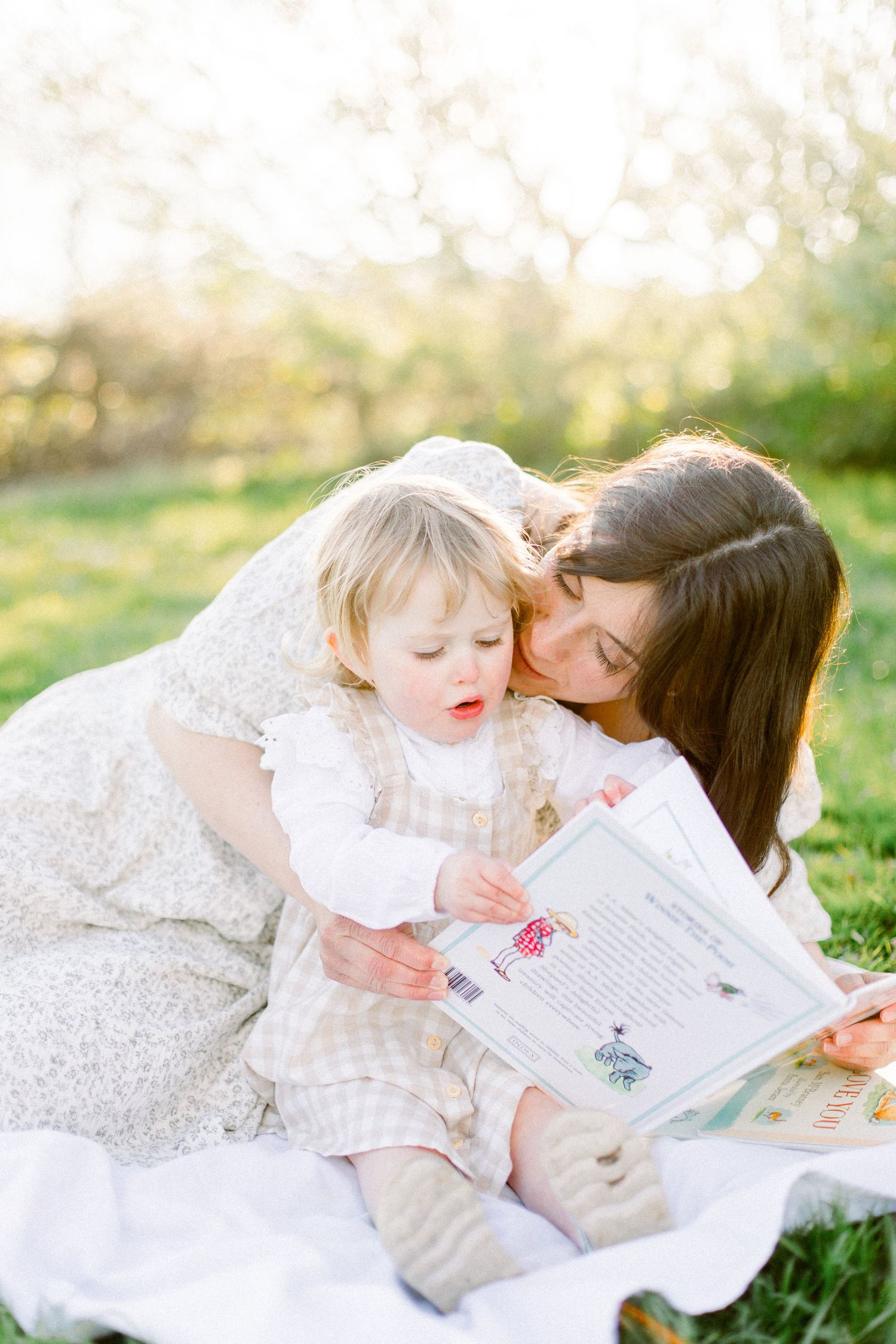 Mother kissing toddler on cheek while reading a book together outdoors in golden sunlight.
