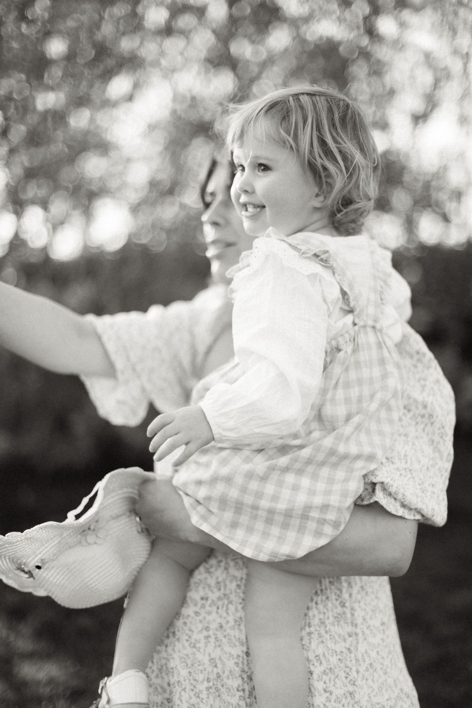 Black and white photo of a smiling toddler girl in a plaid dress being held by a woman outdoors.