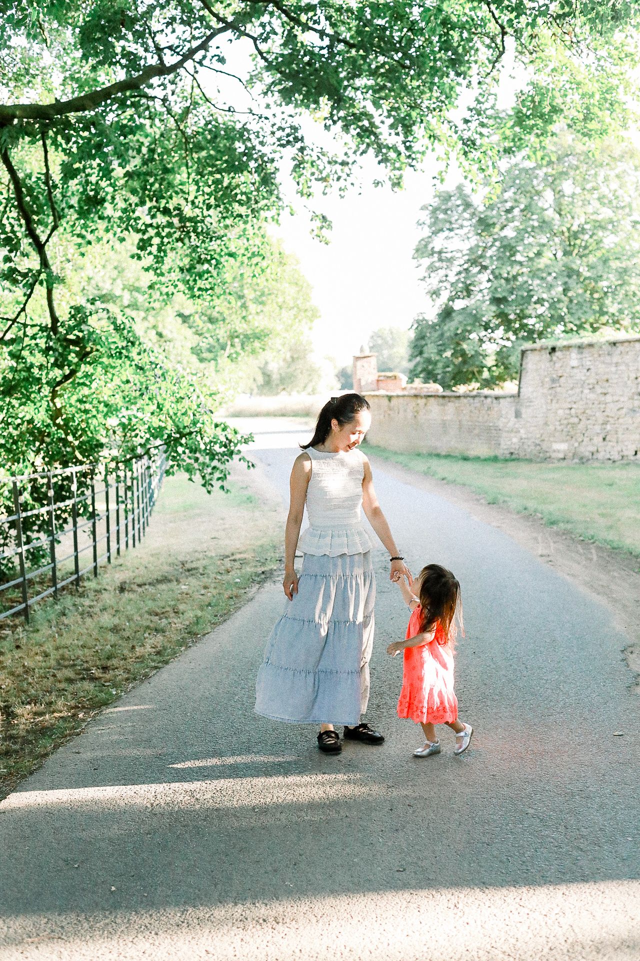 A mother in a blue tiered skirt holds hands with her young daughter in a red dress on a sunlit park path.