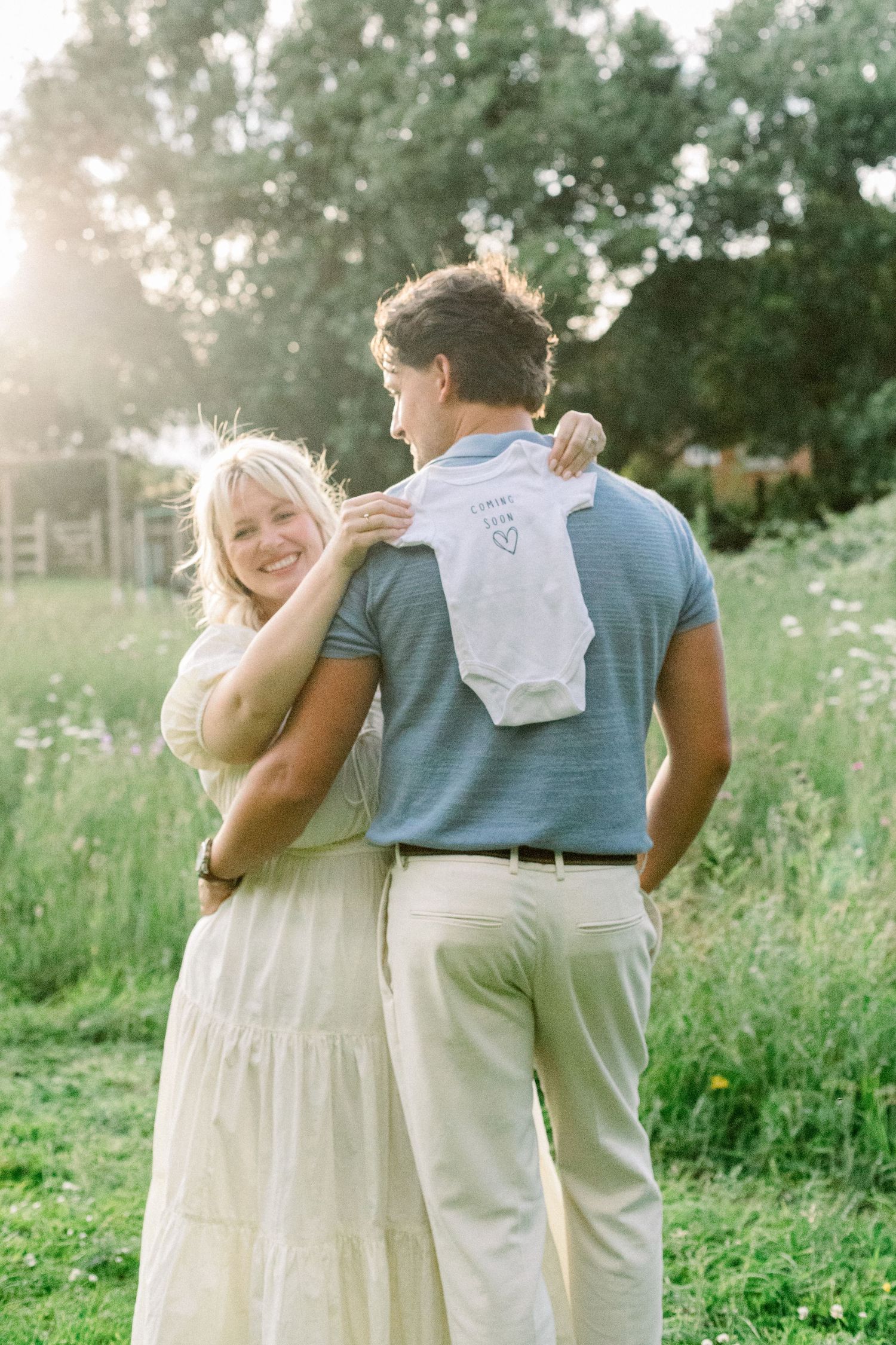 Couple holding a baby onesie in a sunny green field, woman smiling over mans shoulder during pregnancy announcement photo.
