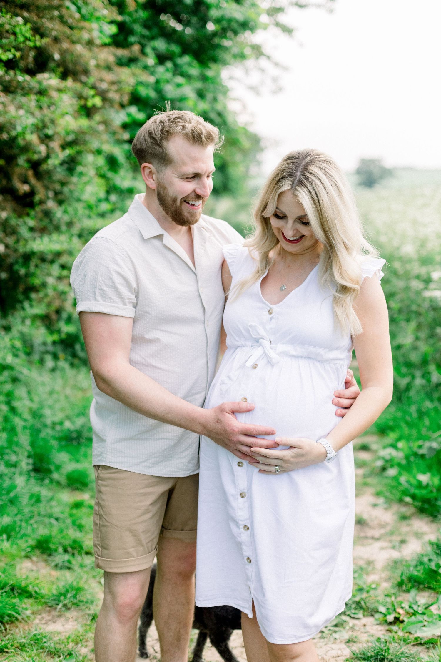 Happy couple holding pregnant belly outdoors in lush green setting, man in beige shirt and woman in white maternity dress.
