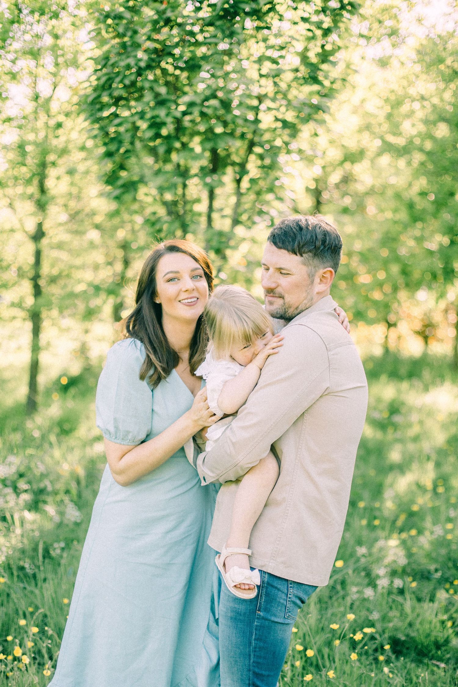 Smiling couple holding their toddler daughter in a sunny green meadow with wildflowers and trees, a warm outdoor family portrait.