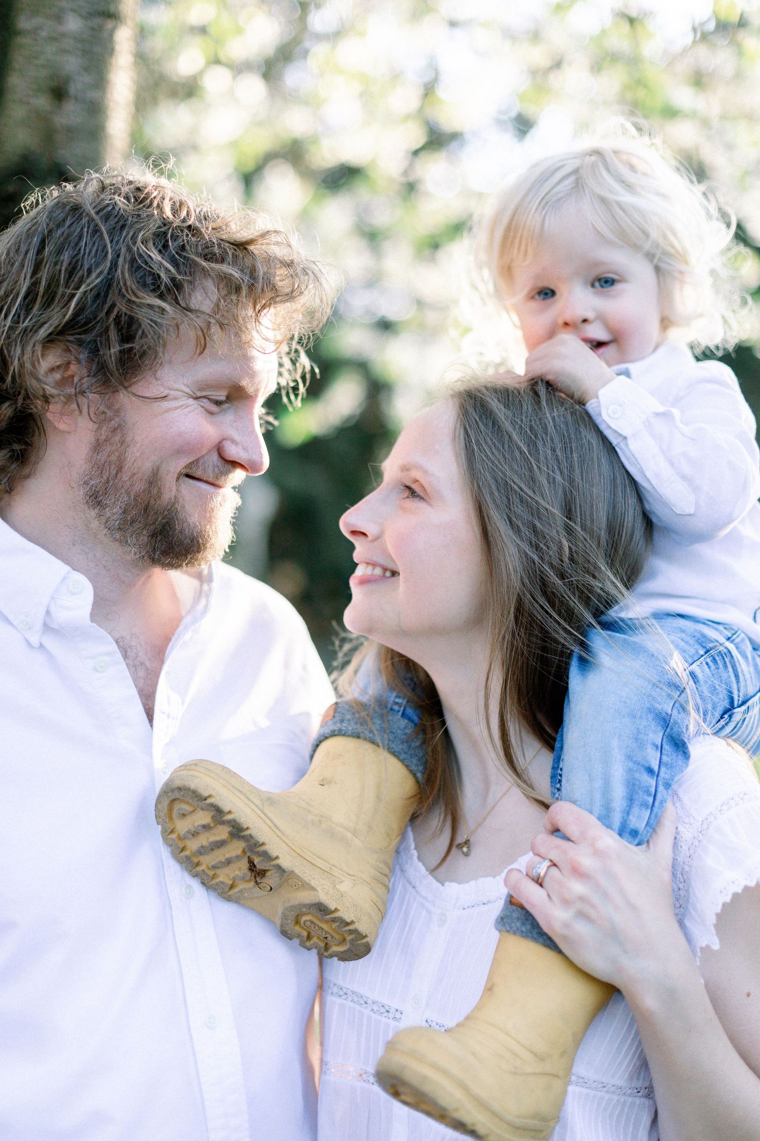 Happy family with curly-haired bearded man, brunette woman, and blonde toddler in yellow boots outdoors.