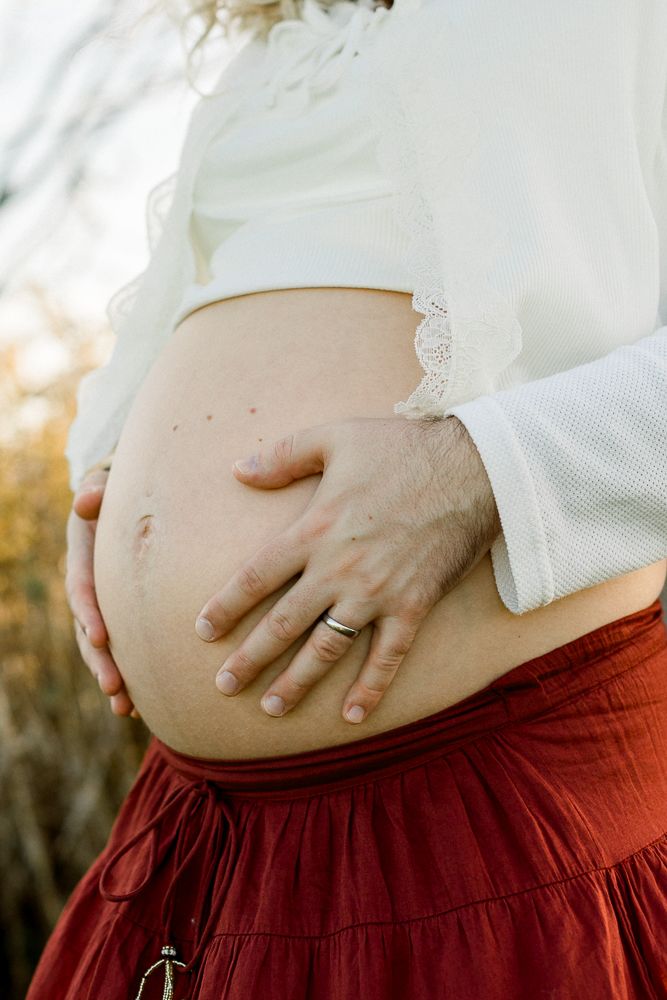 Close-up of a pregnant woman cradling her bare baby bump, wearing a white lace blouse and red skirt outdoors.