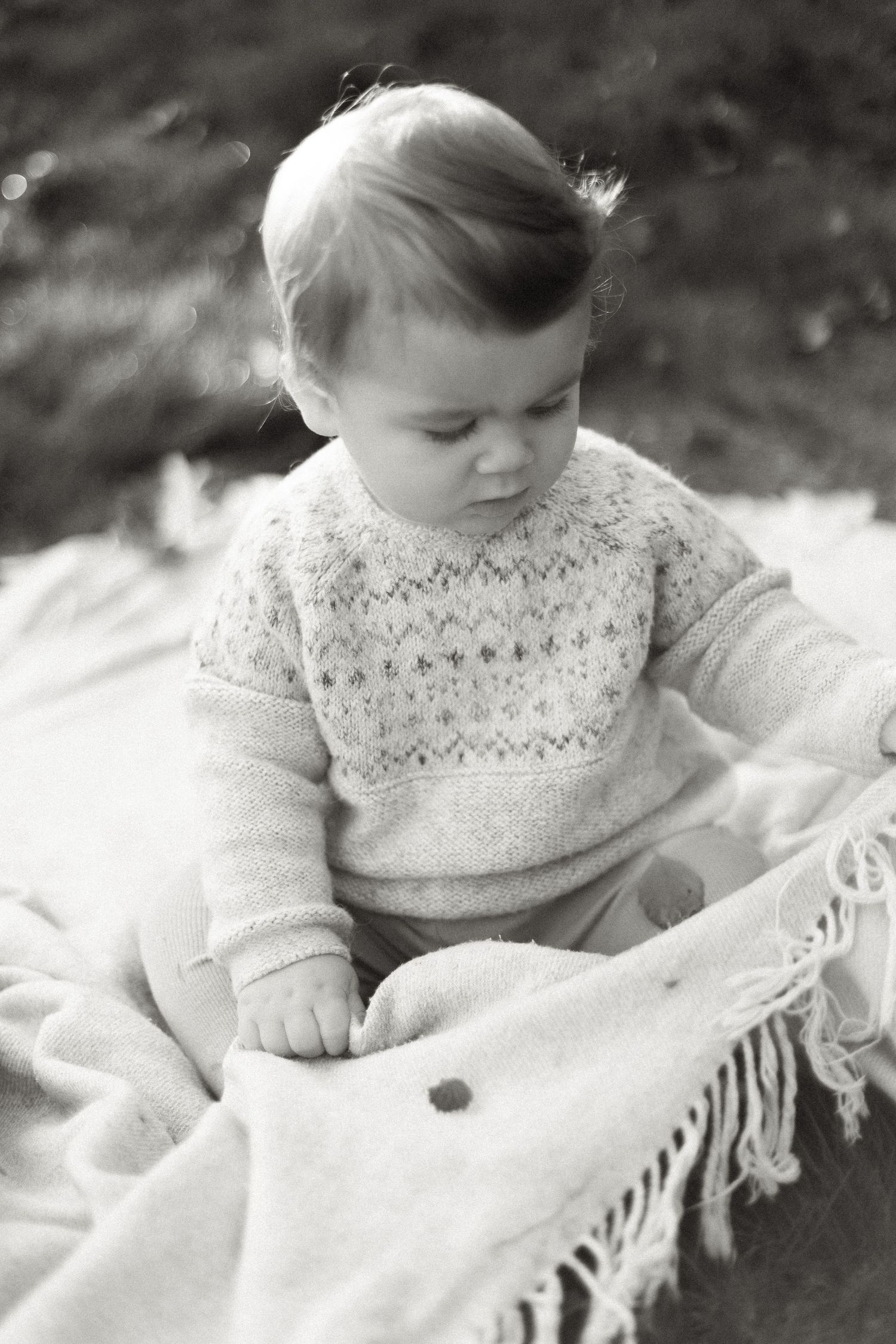 Black and white photo of a baby in a knit sweater sitting on a fringed blanket.
