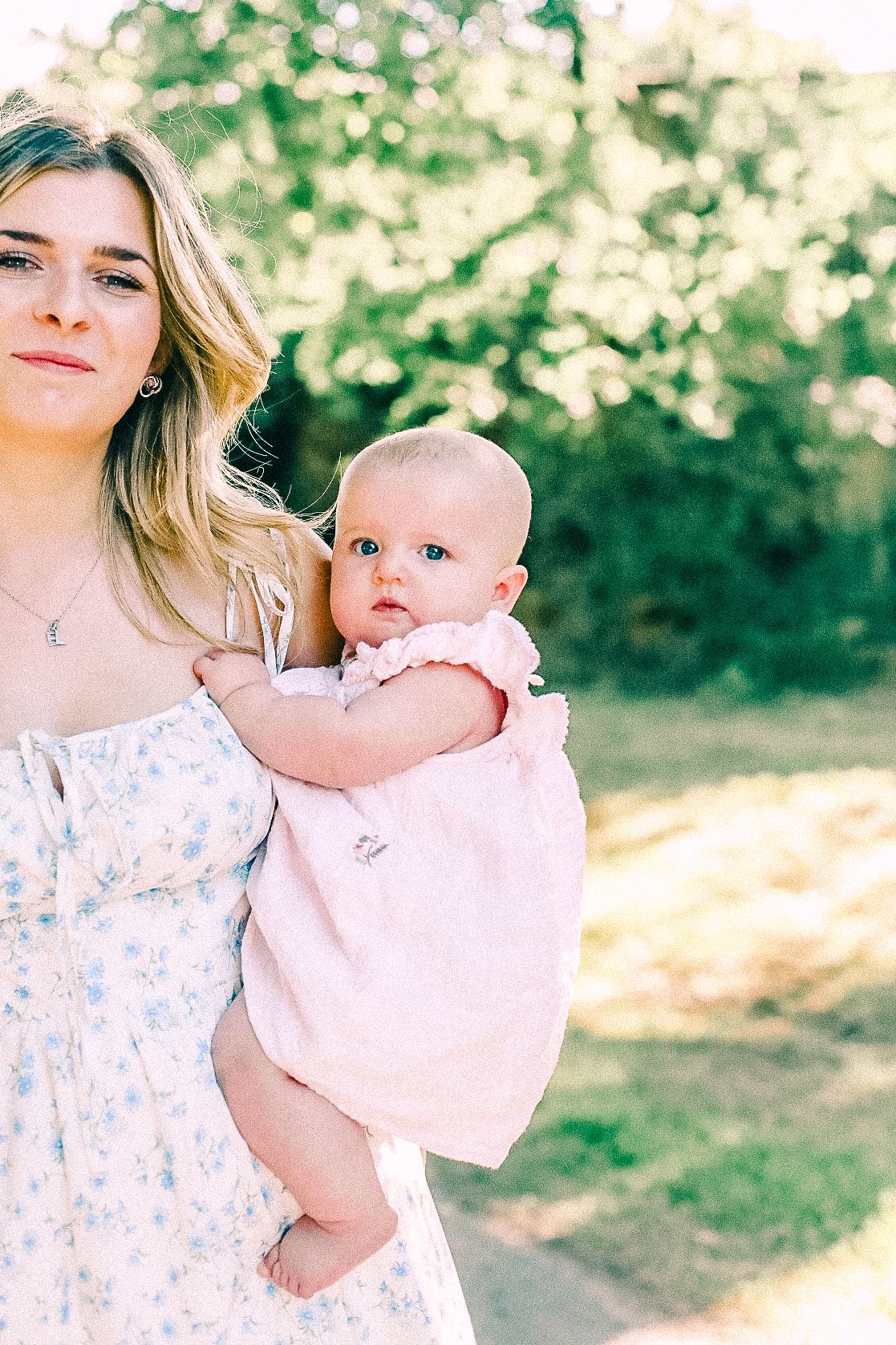 A woman in a floral sundress holds a smiling baby in a pink ruffled outfit outdoors in dappled sunlight with green trees behind.