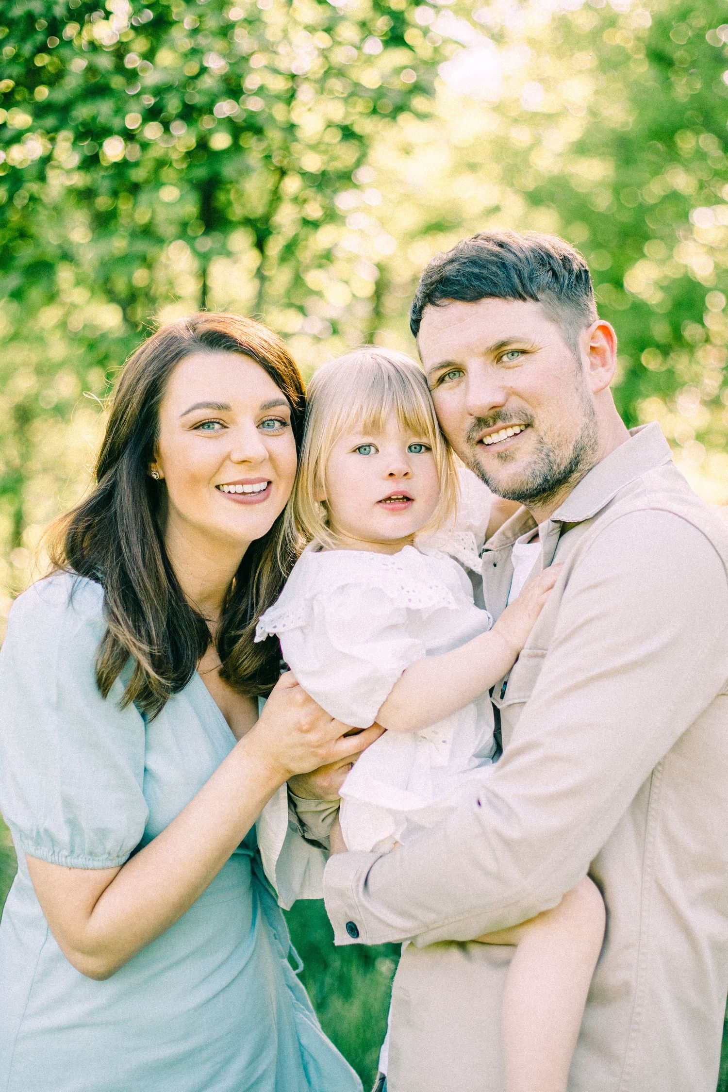 Smiling woman in a blue dress and bearded man in a beige shirt hold a blonde toddler girl outdoors in a sunlit green park.
