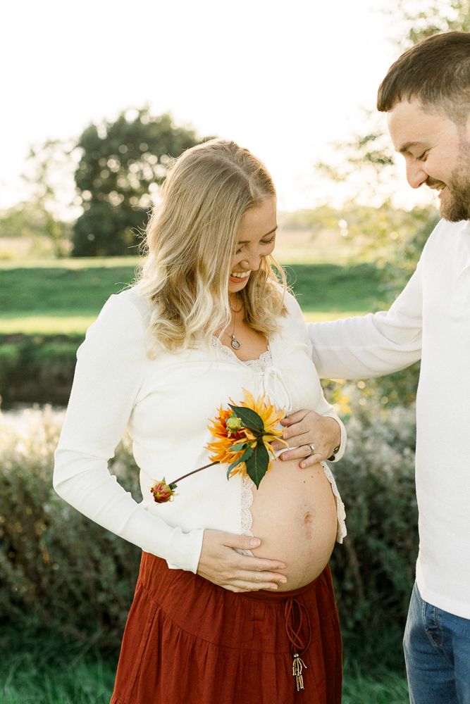 Smiling pregnant woman in white top and red skirt holds sunflower on baby bump with partner outdoors.