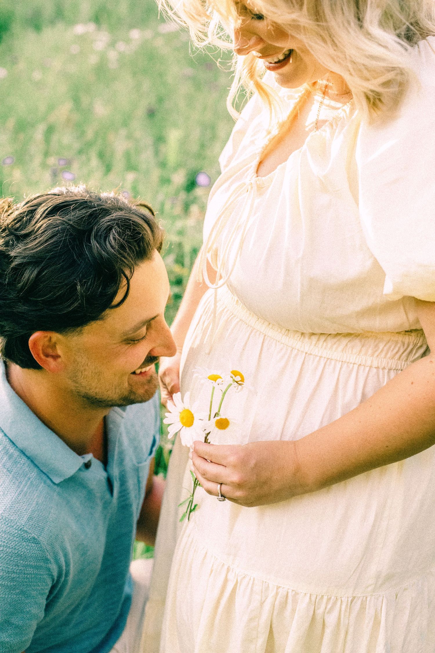 Smiling man kneeling in a wildflower meadow offers a small bouquet of daisies to a pregnant woman in a cream dress.