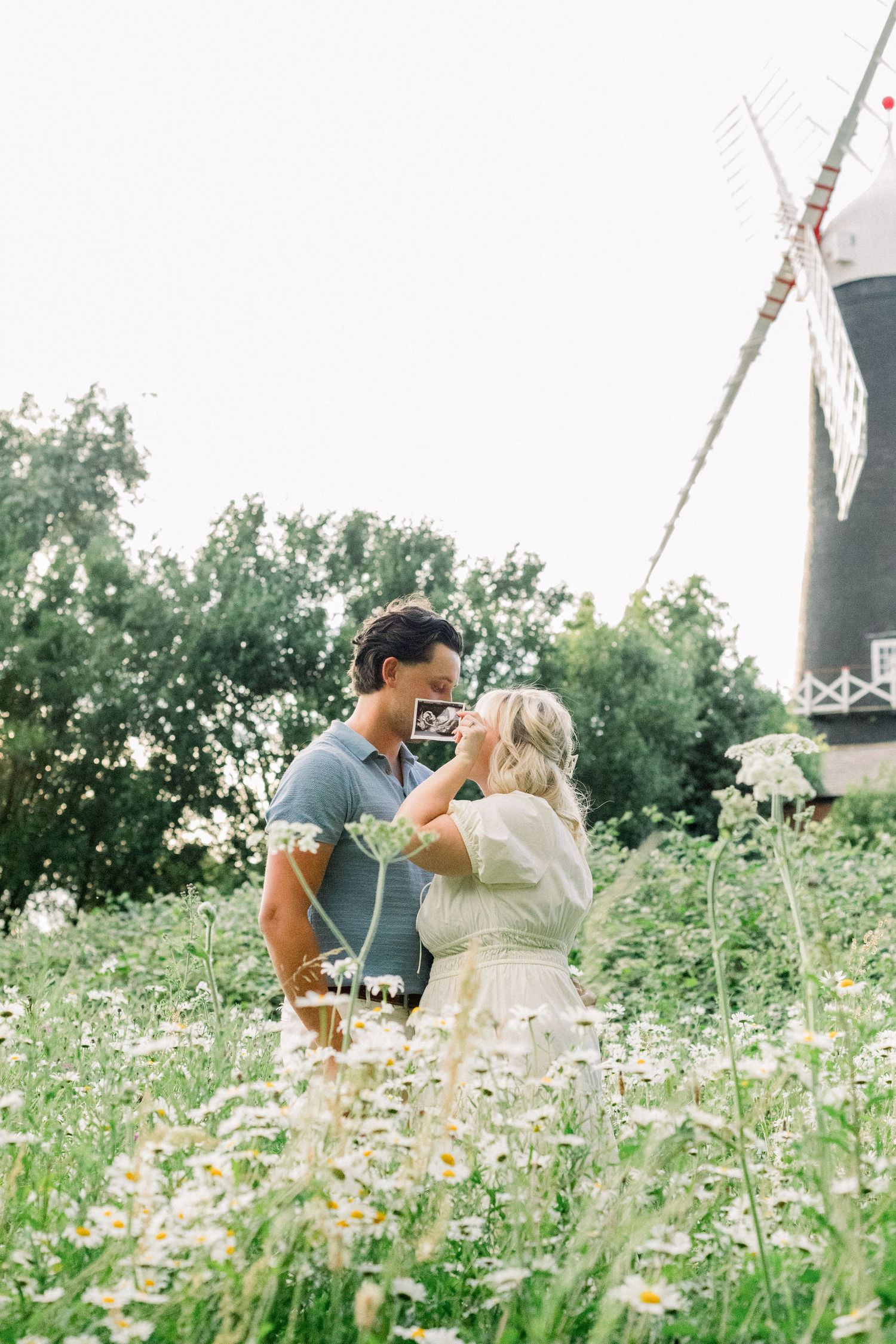 Expectant couple embracing in a field of white daisies holding an ultrasound photo, with a traditional windmill behind them.