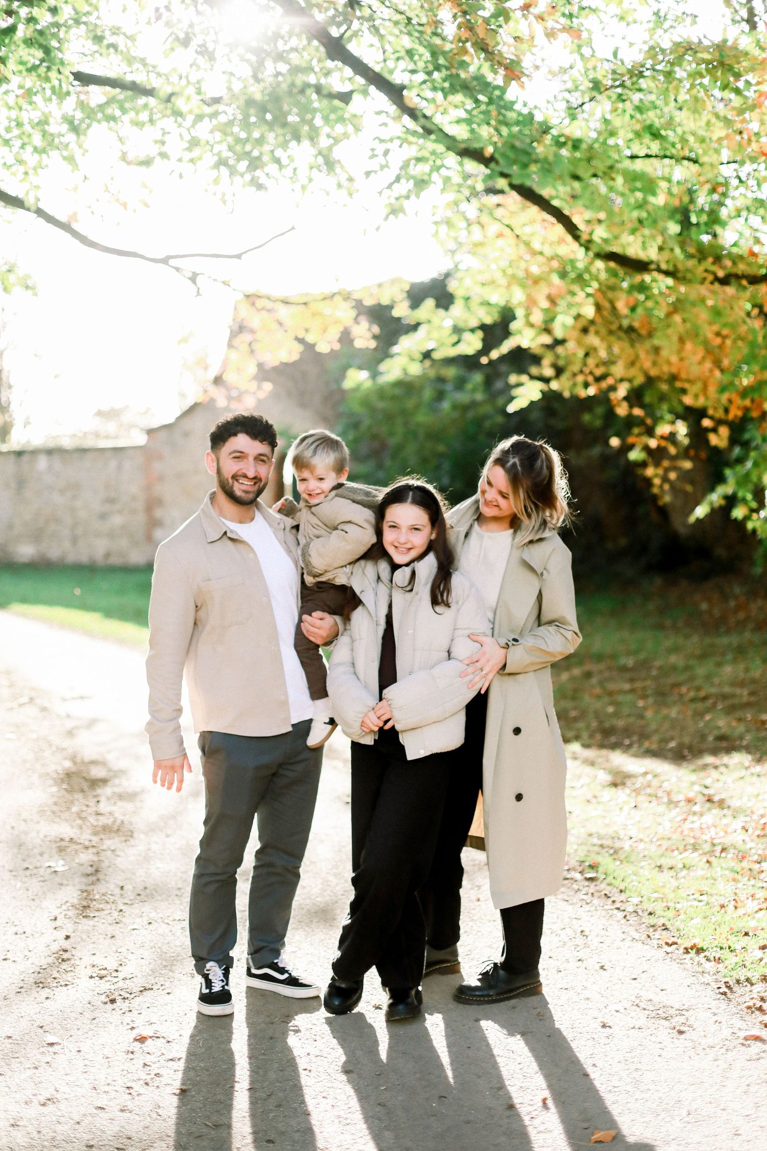 Family of four posing outdoors in autumn, smiling and embracing on a sunlit path surrounded by colorful fall foliage.