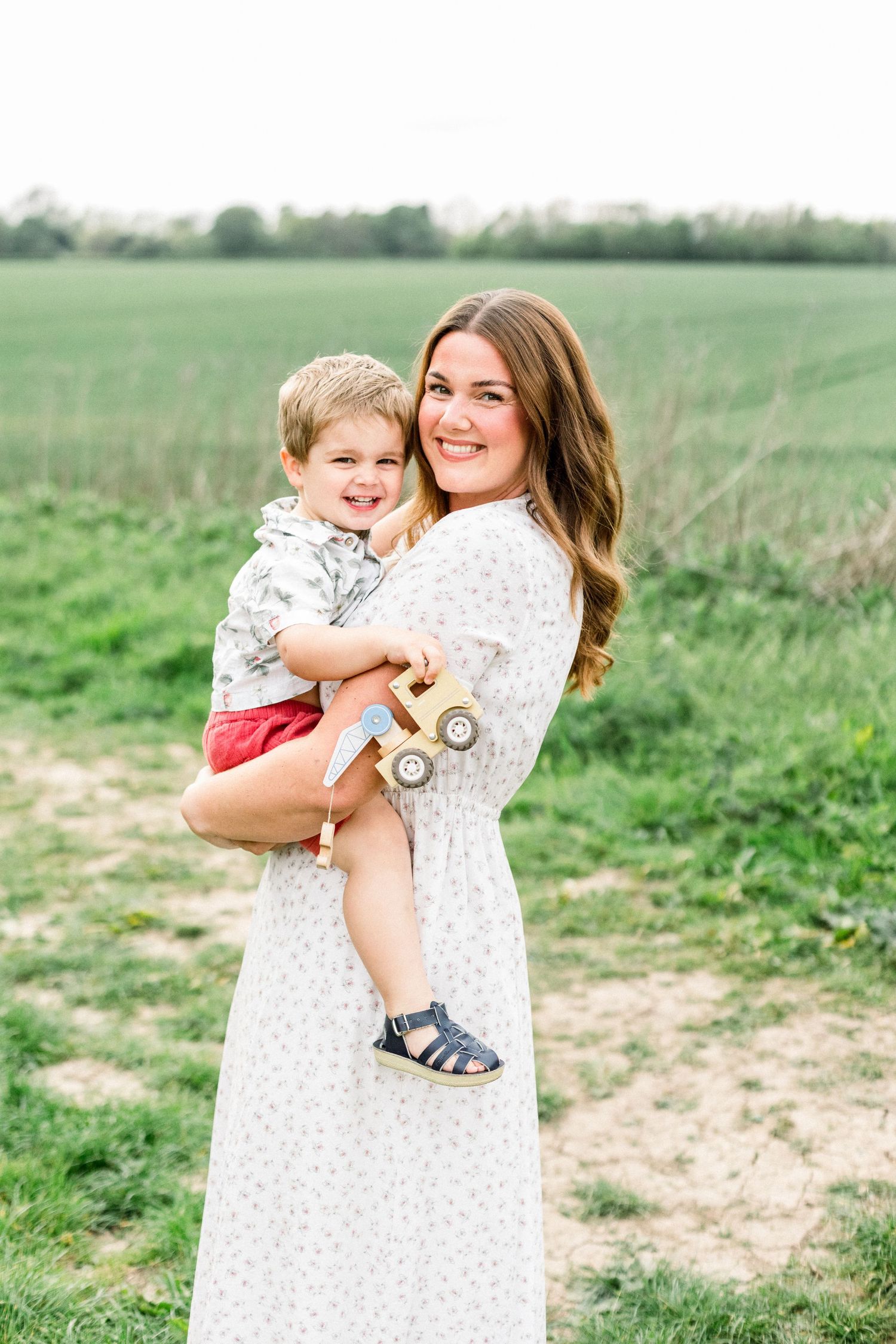 A smiling woman in a floral dress holds a happy toddler boy with a wooden toy truck in a green field.