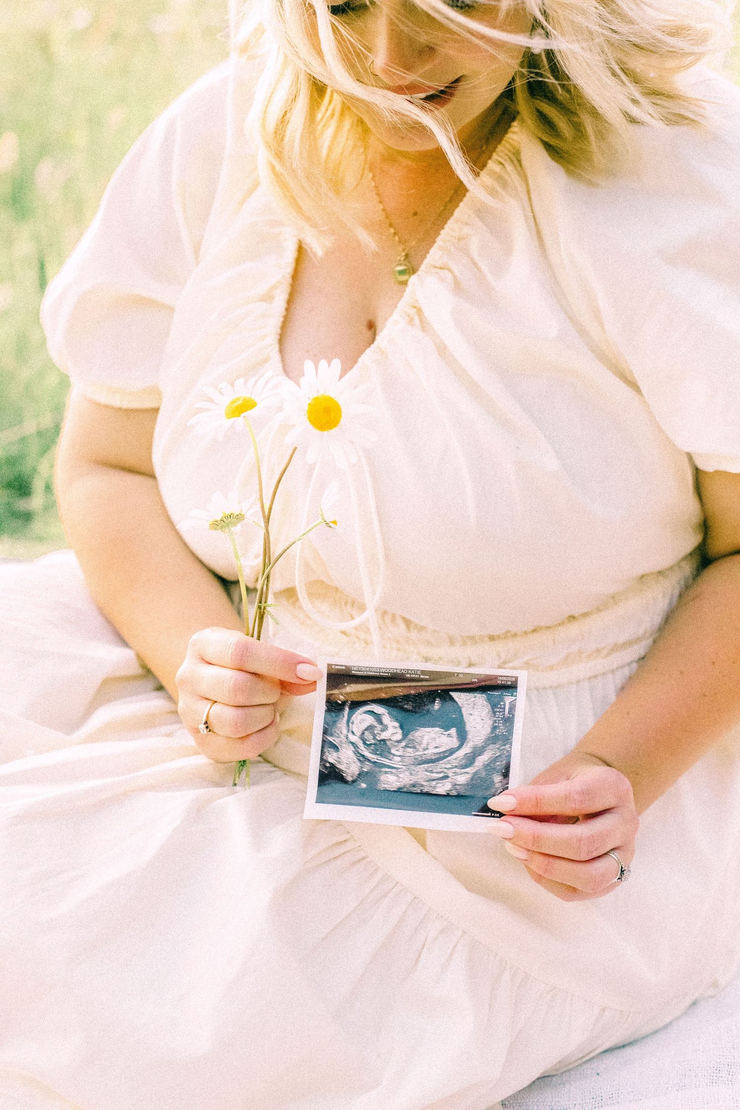 Woman in a cream dress sits outdoors holding an ultrasound photo and daisies, a soft pregnancy announcement portrait.