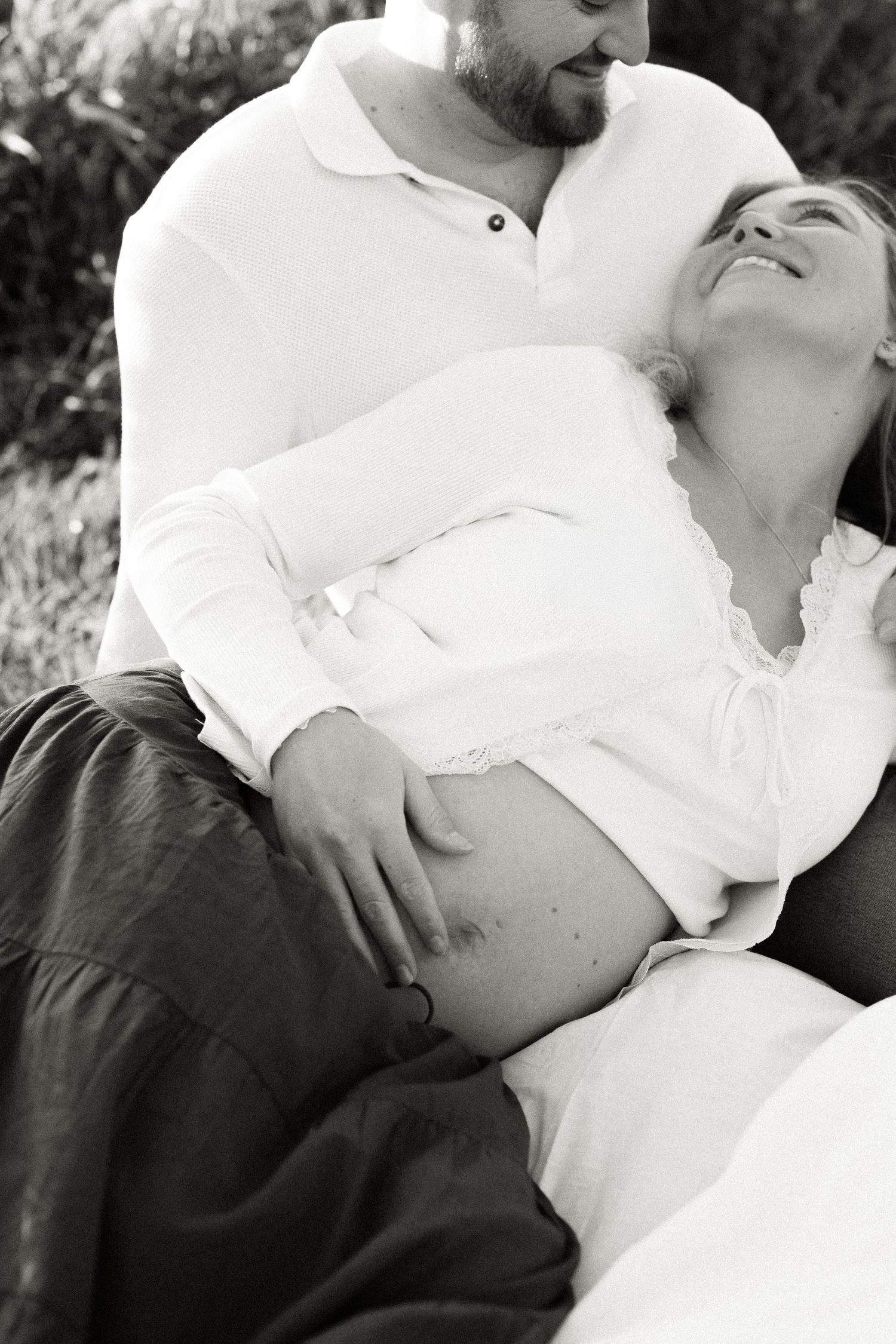 Black and white photo of a couple outdoors, man embracing pregnant woman as she reclines against him.