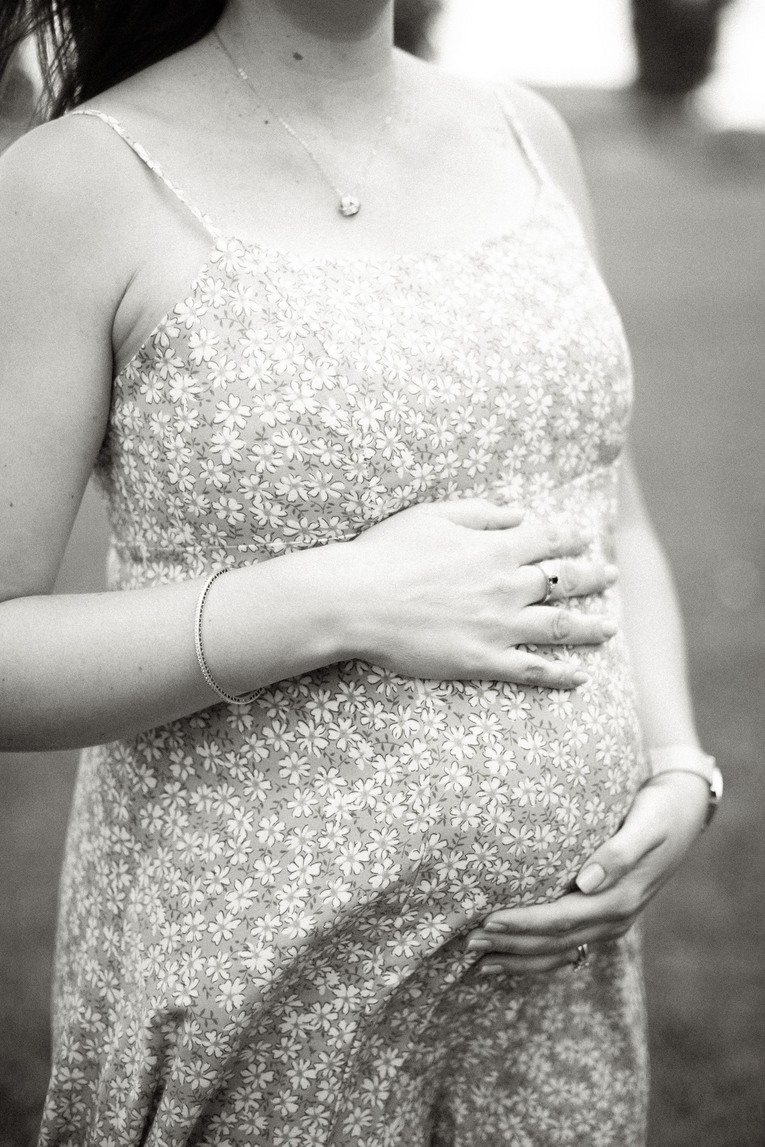 Black and white photo of a pregnant woman in a floral dress cradling her baby bump with both hands.