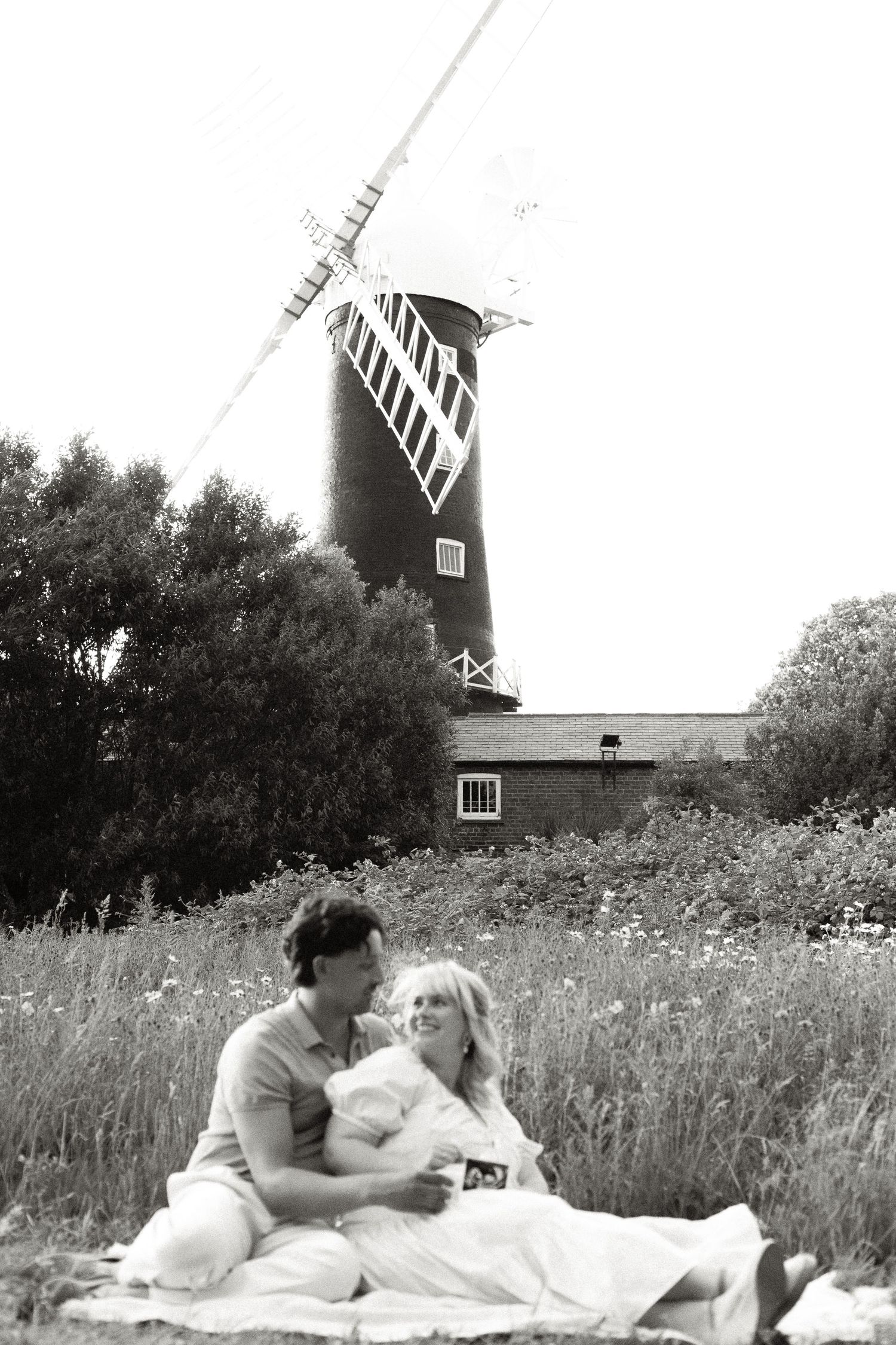 Black and white photo of a couple sitting on a picnic blanket in a wildflower meadow with a historic windmill in the background.
