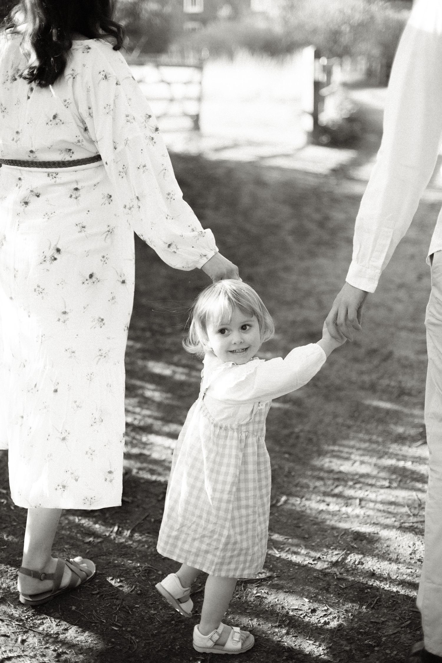 Black and white image of a child in a gingham dress holding hands with adults on a sunny path.