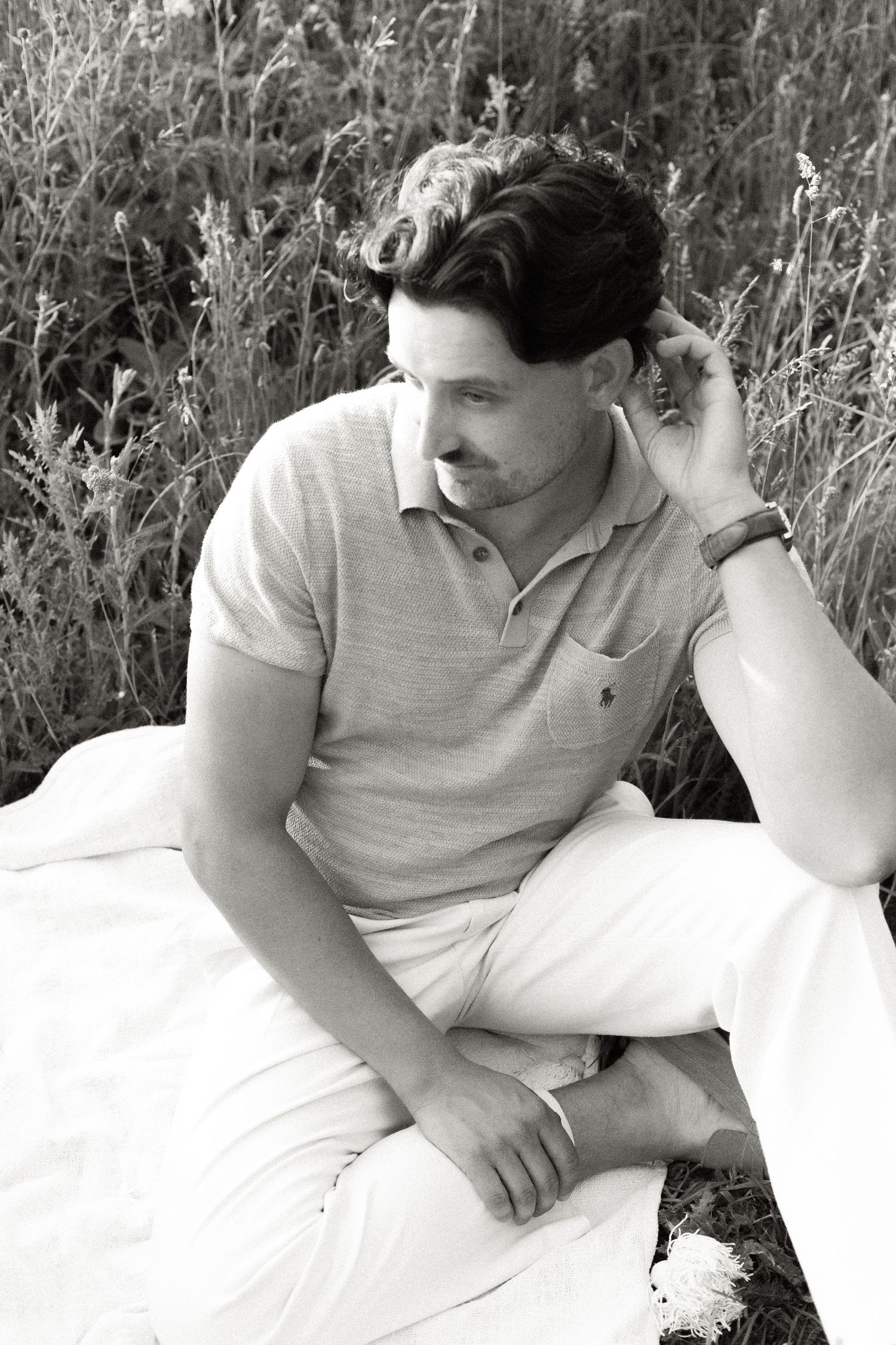 Black and white photo of a man in a polo shirt and light trousers sitting on a blanket in tall grass, resting his hand against his head.