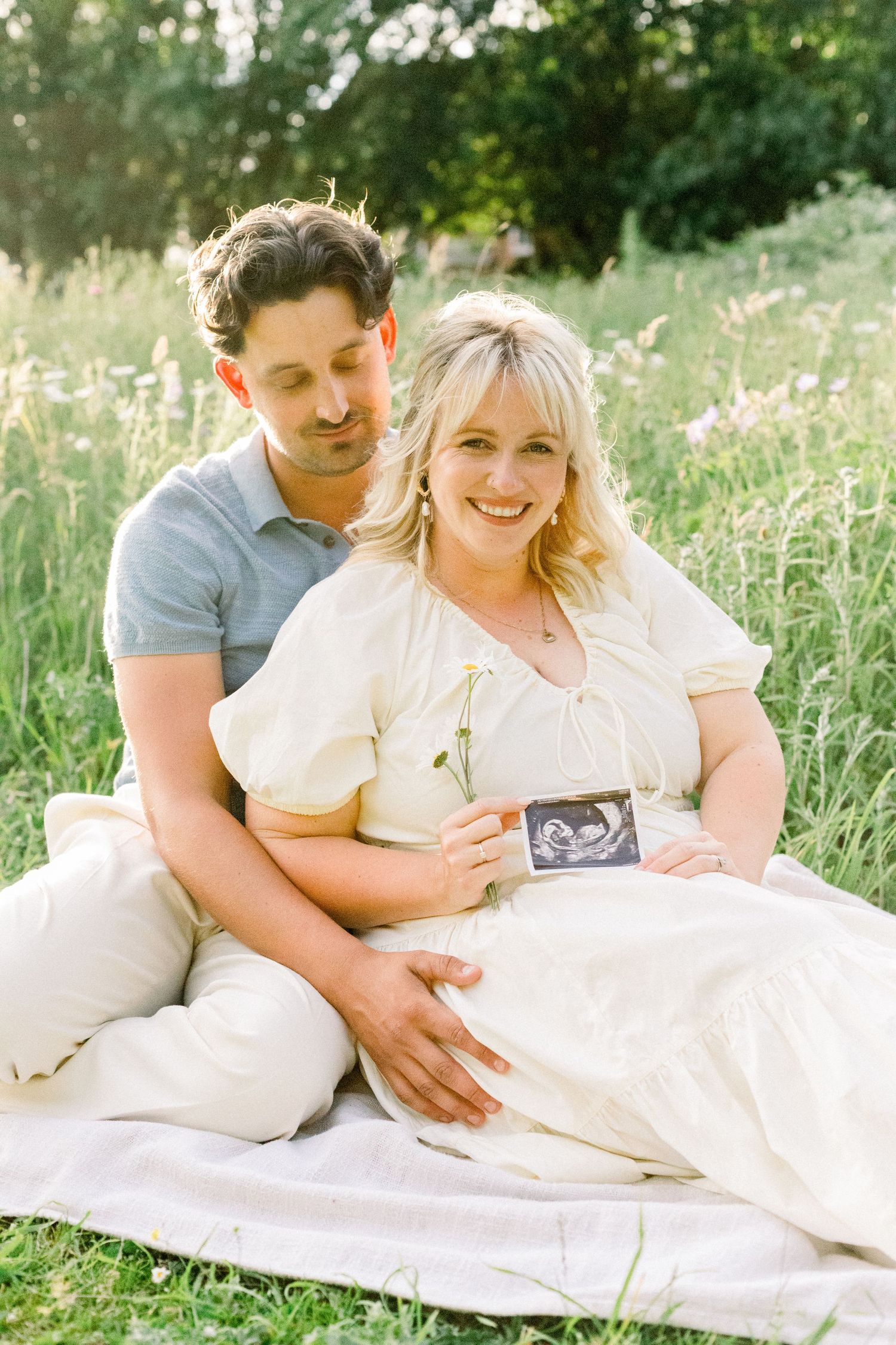 Happy pregnant couple sitting on blanket in meadow, holding ultrasound photo, smiling outdoors in golden sunlight.