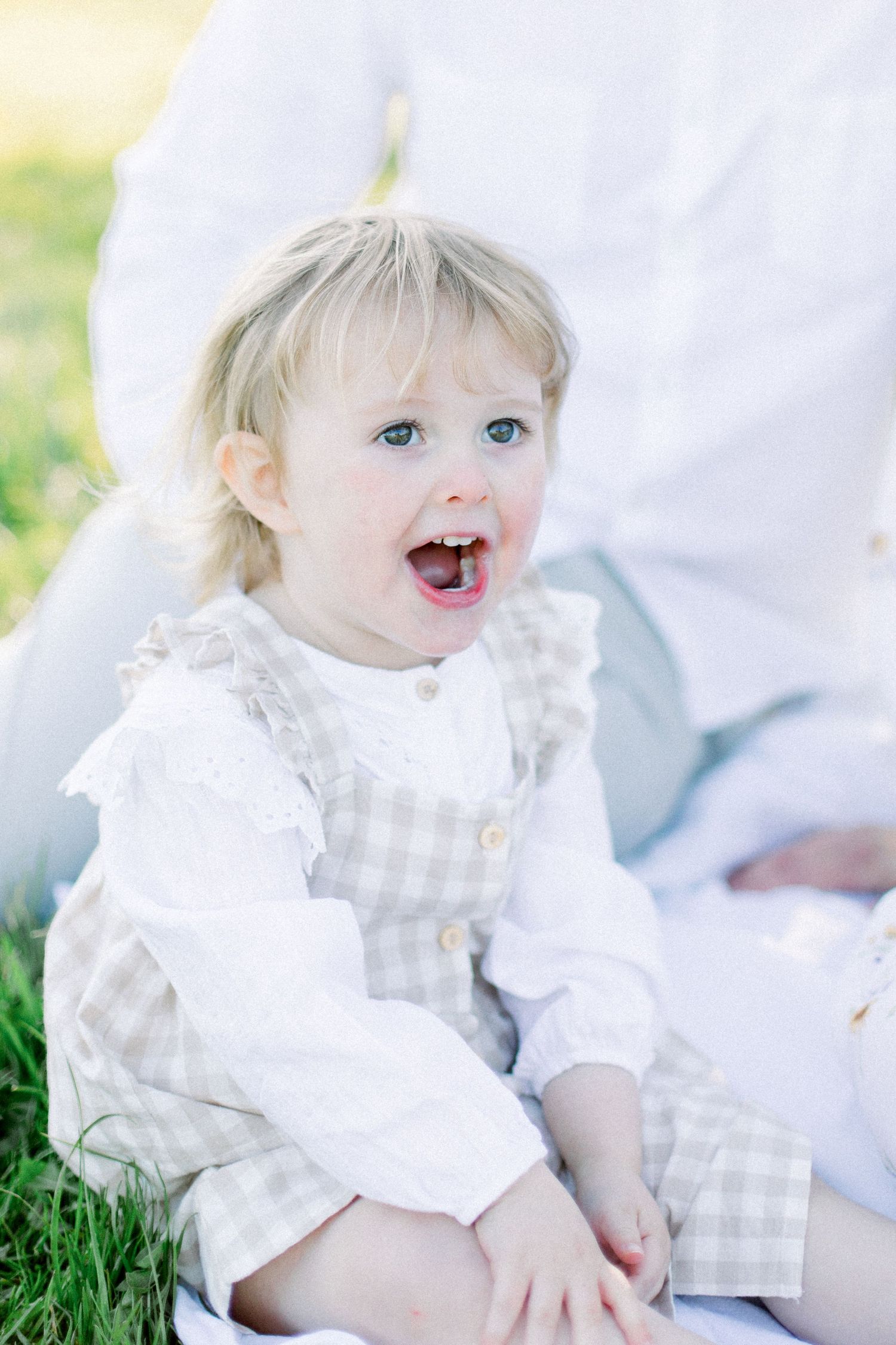 An adorable blonde toddler girl in a gingham dress sits on a white blanket in a sunny green garden.