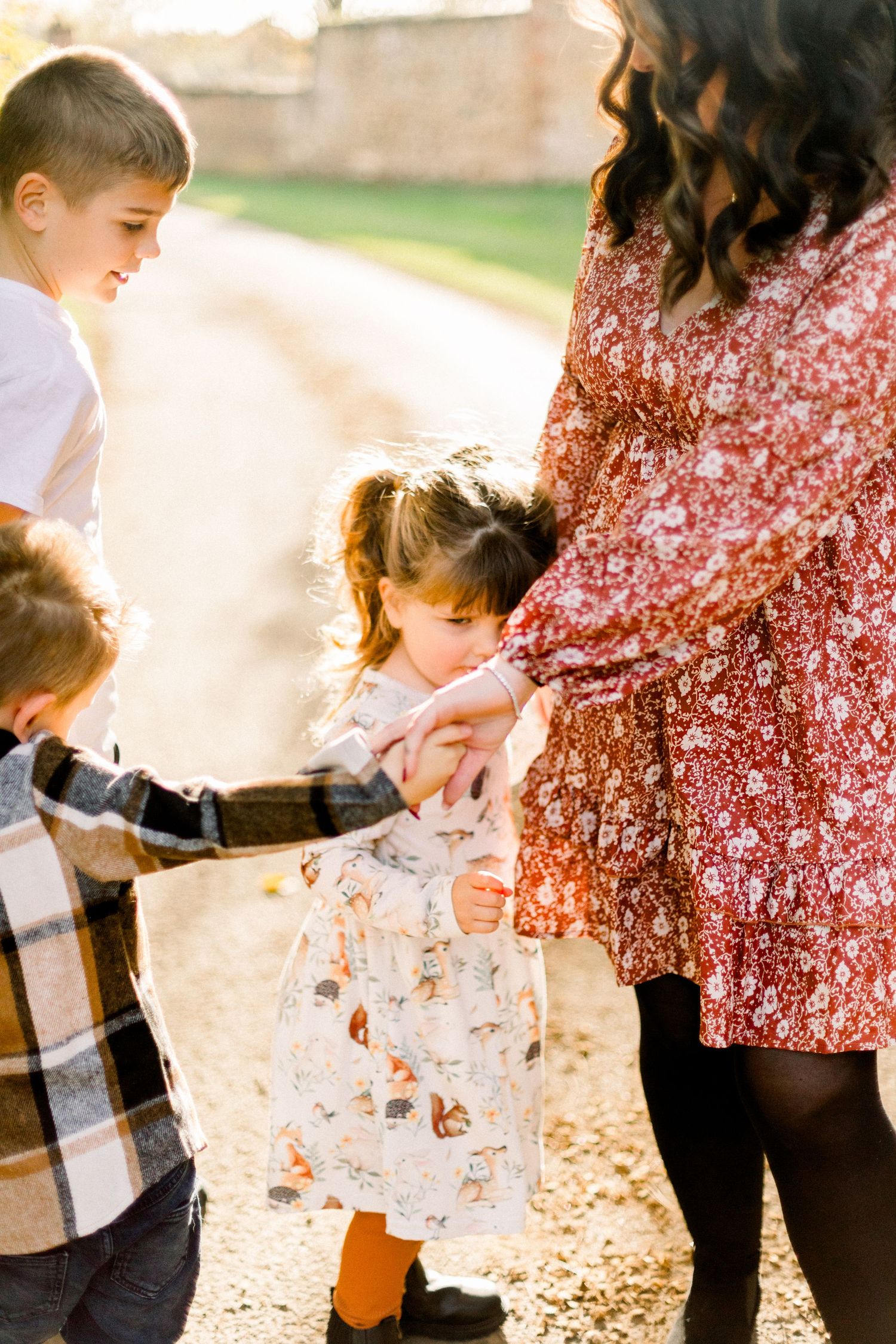 A mother in a floral dress holds hands with her young children outdoors in warm golden light.