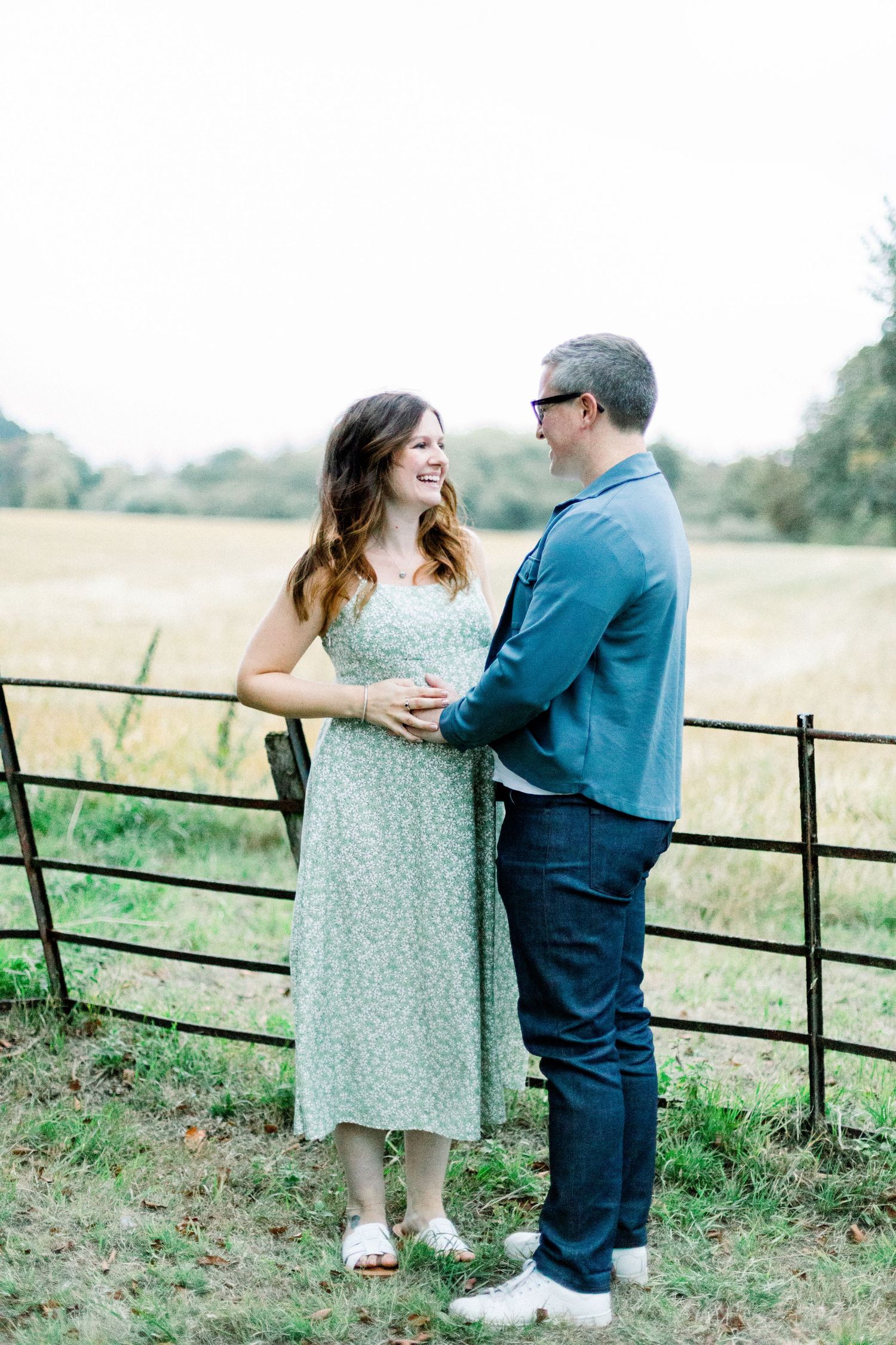 A couple smiles at each other holding hands by a rustic fence in a green field, woman in floral dress, man in blue blazer.