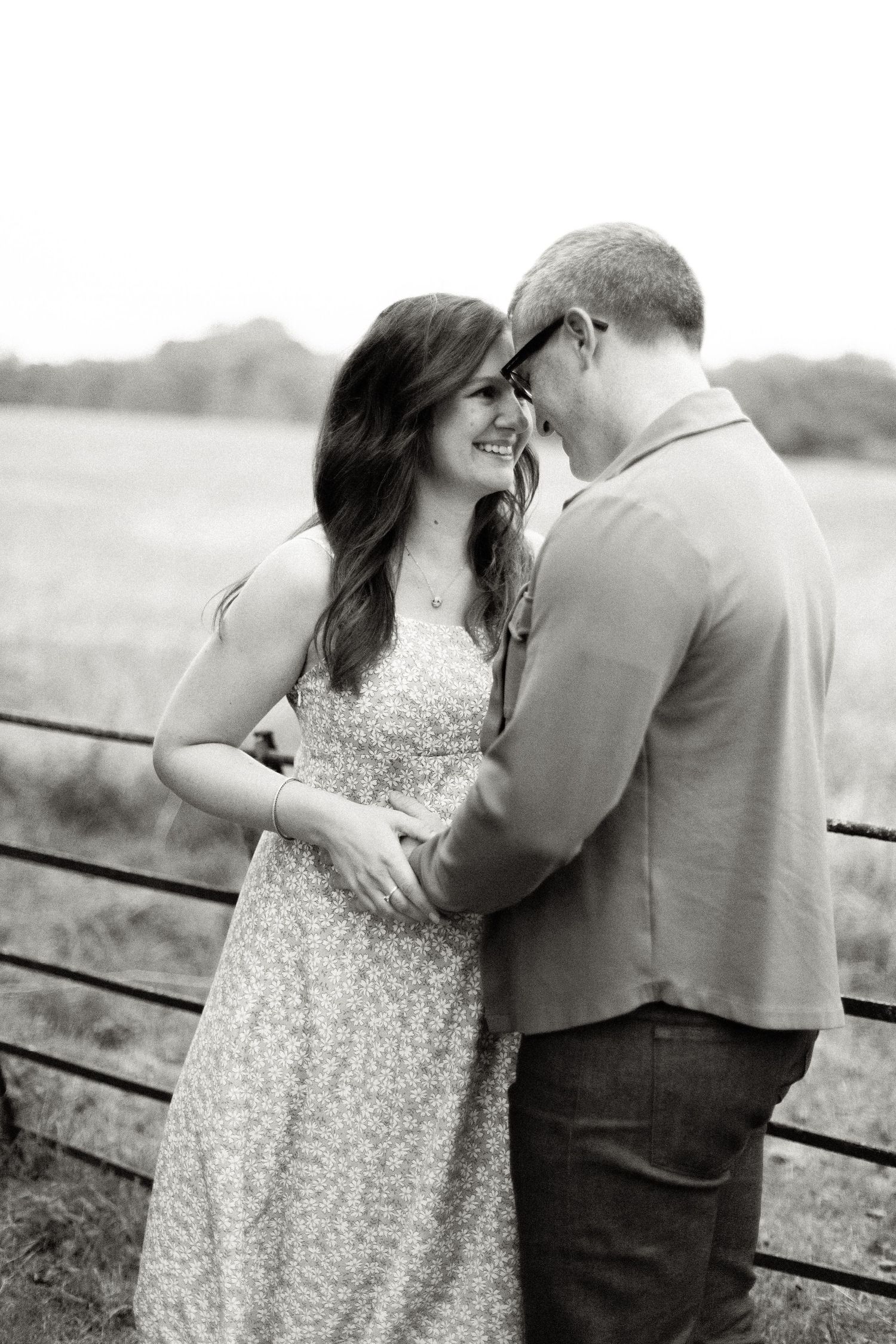 Black and white photo of a smiling couple embracing near a fence in an open field during an outdoor engagement session.