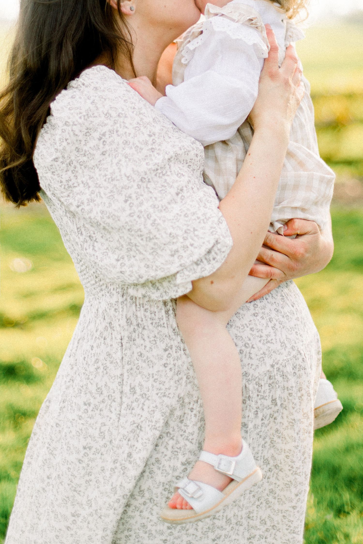 Woman in a floral dress holds and kisses a toddler in white sandals while standing in a sunlit green field.