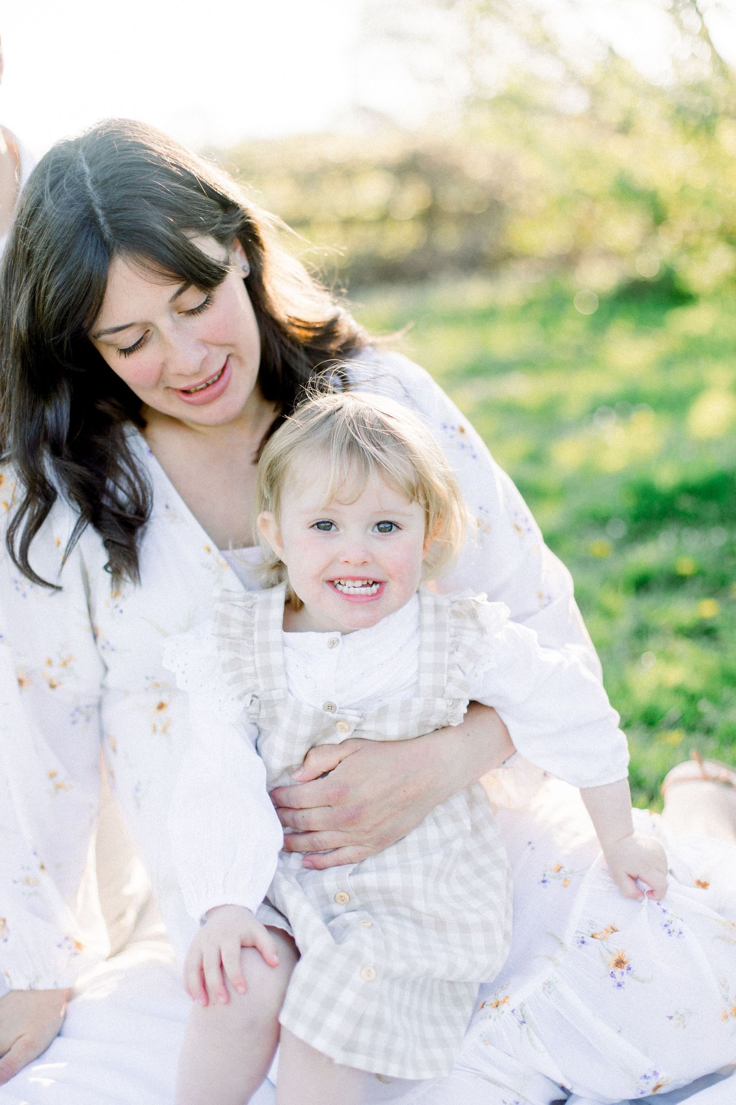 A dark-haired mother in a floral dress smiles down at her blonde toddler daughter sitting on her lap outdoors.