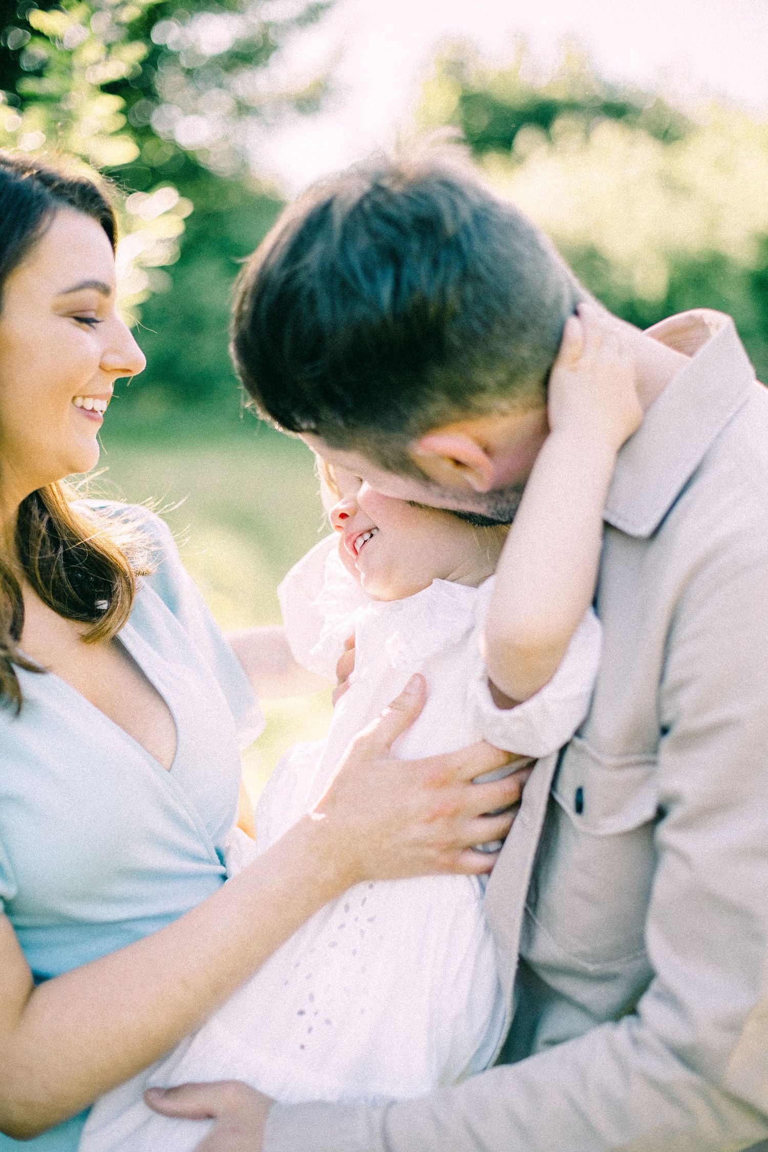 Smiling woman and man in soft sunlight cuddle a laughing toddler in a white dress during an outdoor family portrait session.