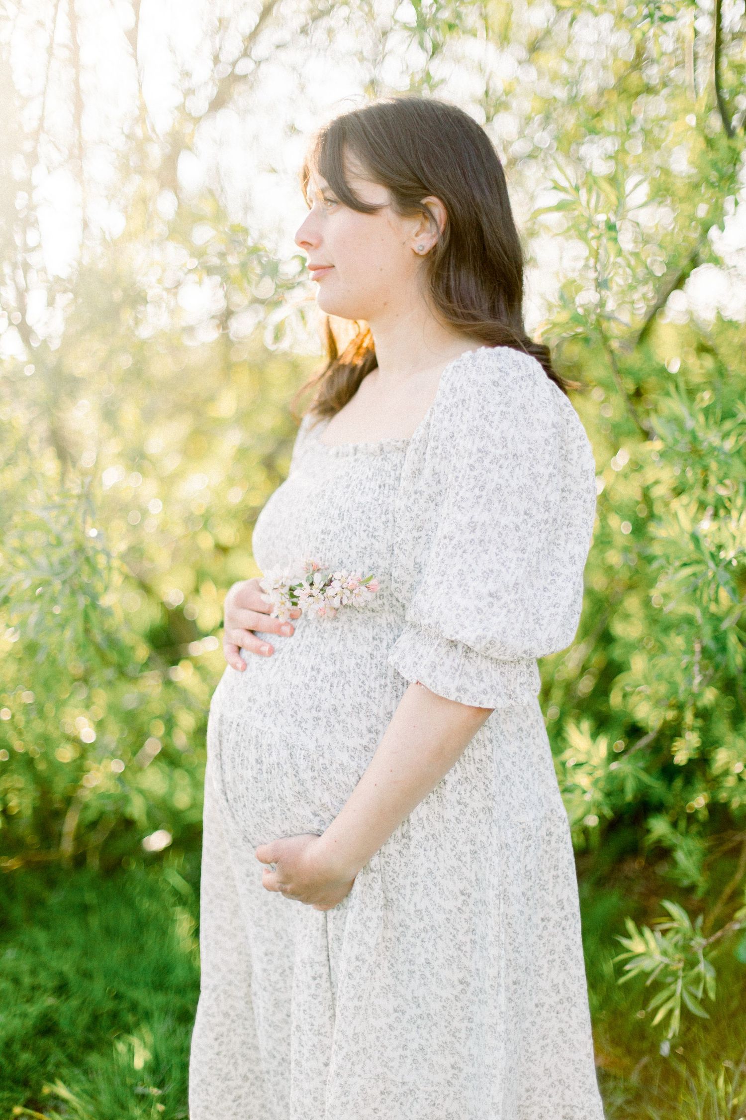 A woman in a white dress stands in profile against sunlit foliage while touching her pregnant belly.