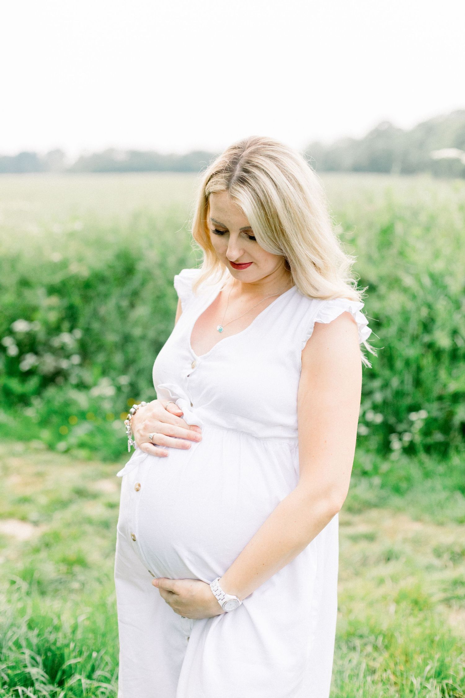 Pregnant woman in white dress cradling baby bump in a green field, looking down with a gentle smile.